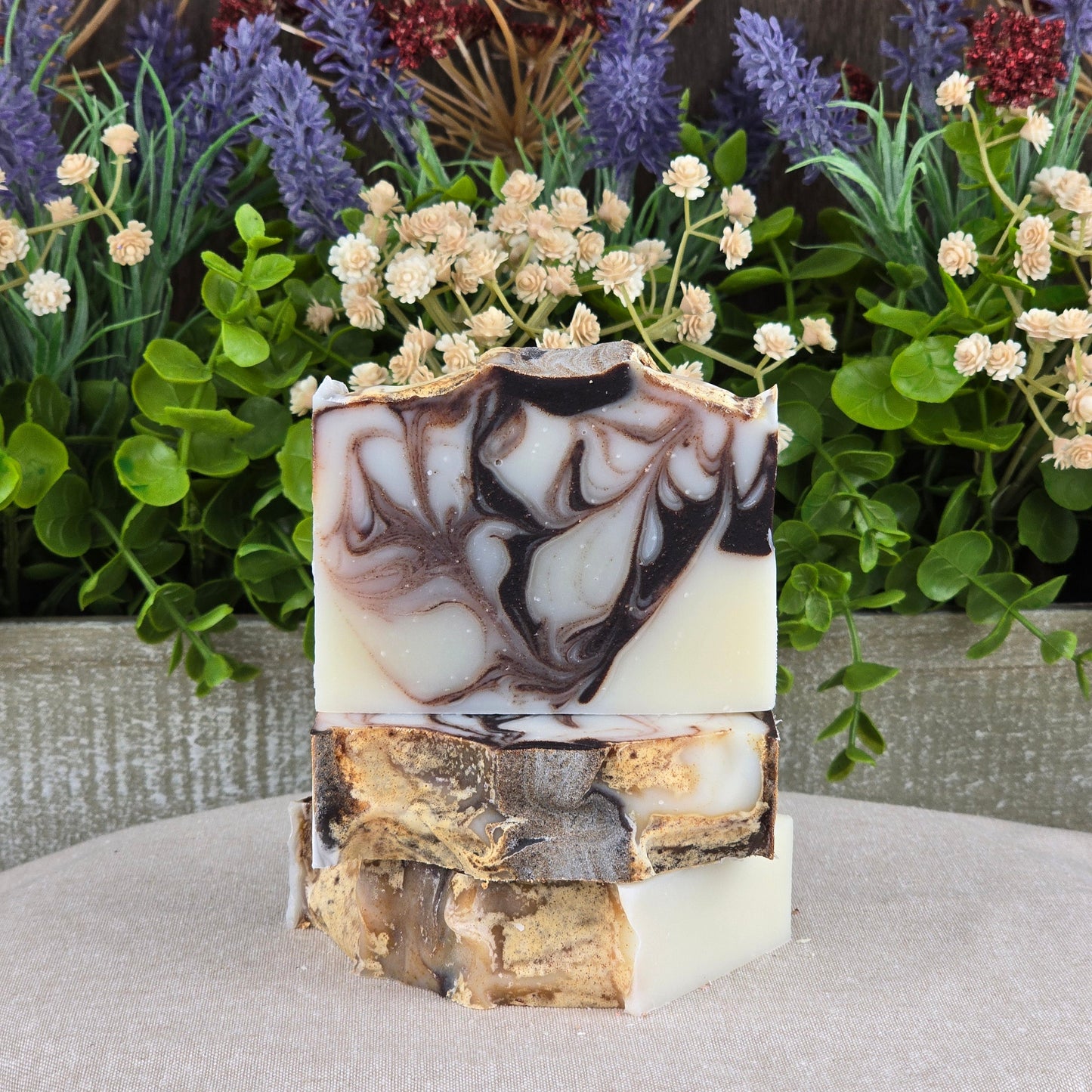 Three Cedarwood + Tallow Handmade Artisan Soap bars by Jarhead Aromatics, featuring natural brown, white, and cream swirls, are stacked on a neutral beige surface with green foliage and purple flowers in the background.