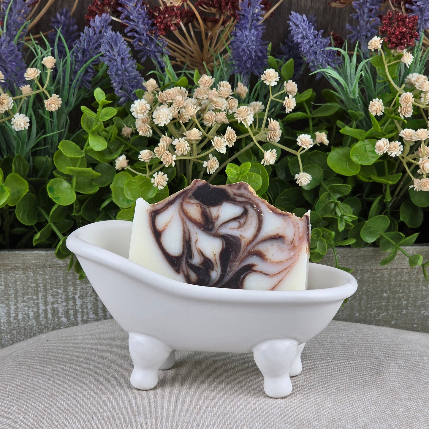 Jarhead Aromaticsโ Cedarwood + Tallow Handmade Artisan Soap, featuring a brown and white marbled pattern, sits in a white clawfoot bathtub dish with green leaves and faux white and purple flowers as a charming backdrop.