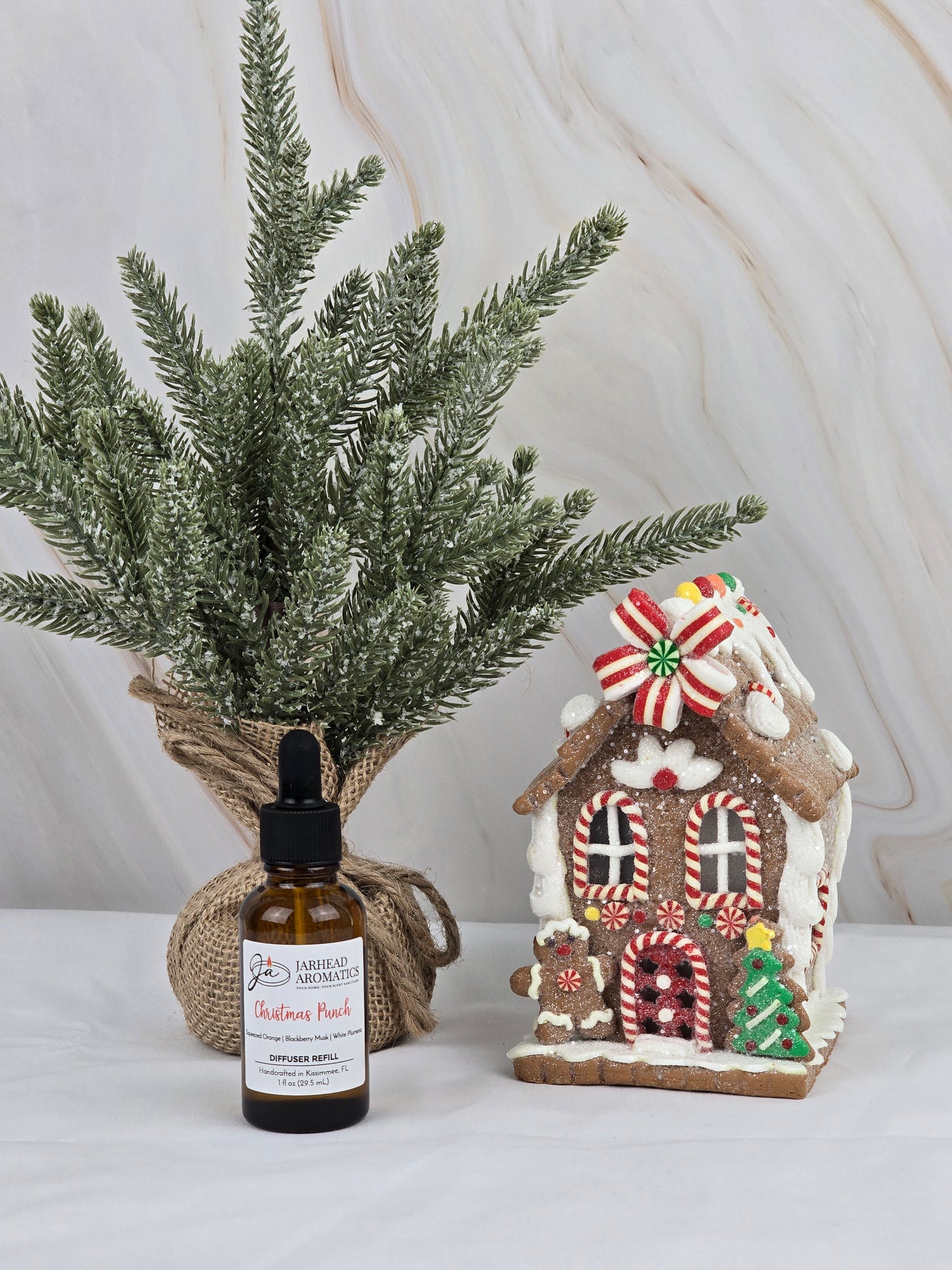 The Christmas Punch Car Diffuser Refill from Jarhead Aromatics emits a festive scent as it sits beside a small, decorated gingerbread house and a miniature burlap-wrapped fir tree against a marble backdrop.