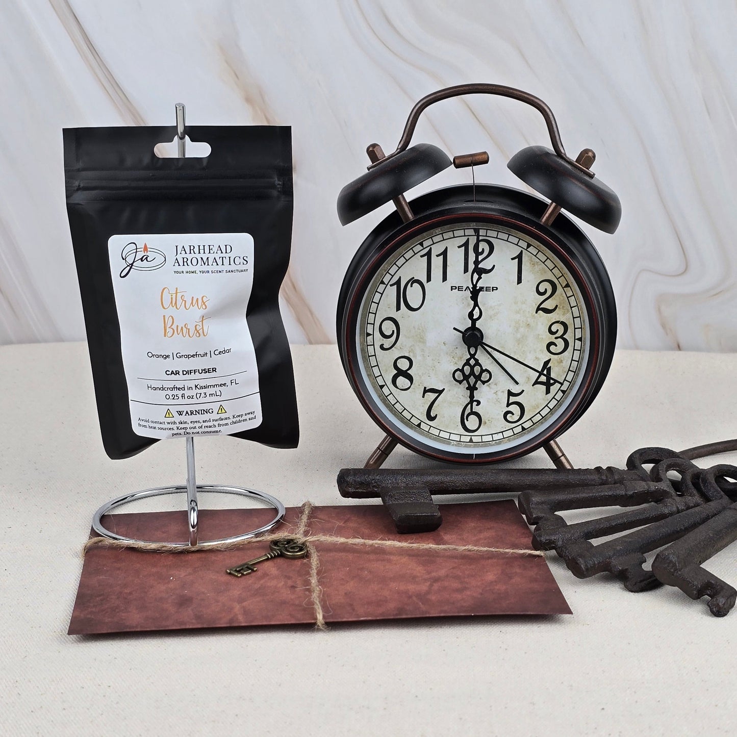 A black vintage alarm clock, Jarhead Aromatics’ Citrus Burst - Car Diffuser, an old-fashioned key on a brown envelope, and several large iron keys are arranged on a cream surface with a light marble background.