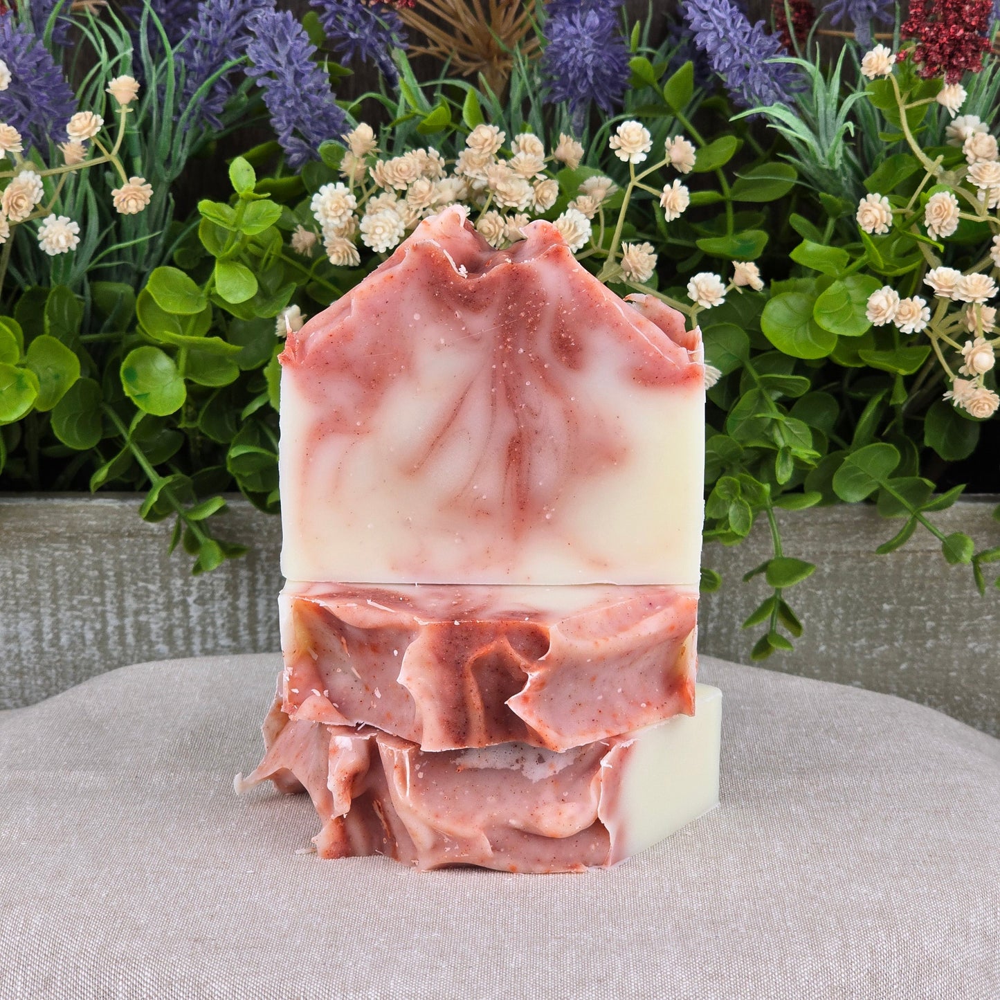 Two Citrus + Tallow Handmade Artisan Soap bars by Jarhead Aromatics, with pink and white marbled swirls and ruffled tops, are stacked before green leaves and tiny white and purple flowers. Made with grass-fed beef tallow for a natural, unique look.