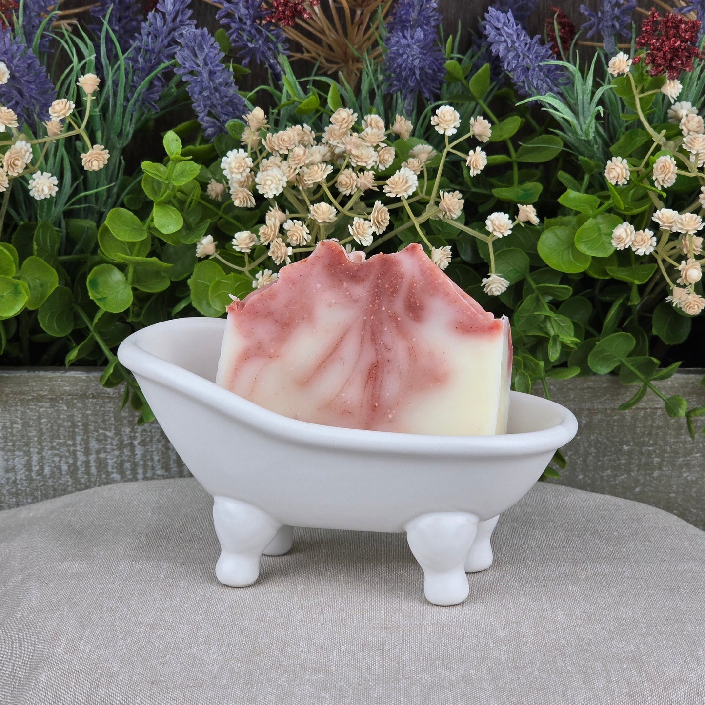 The Citrus + Tallow Handmade Artisan Soap by Jarhead Aromatics features pink and white swirls, displayed in a white clawfoot tub soap dish with greenery and purple flowers. Made with nourishing grass-fed beef tallow for a luxurious, all-natural cleanse.
