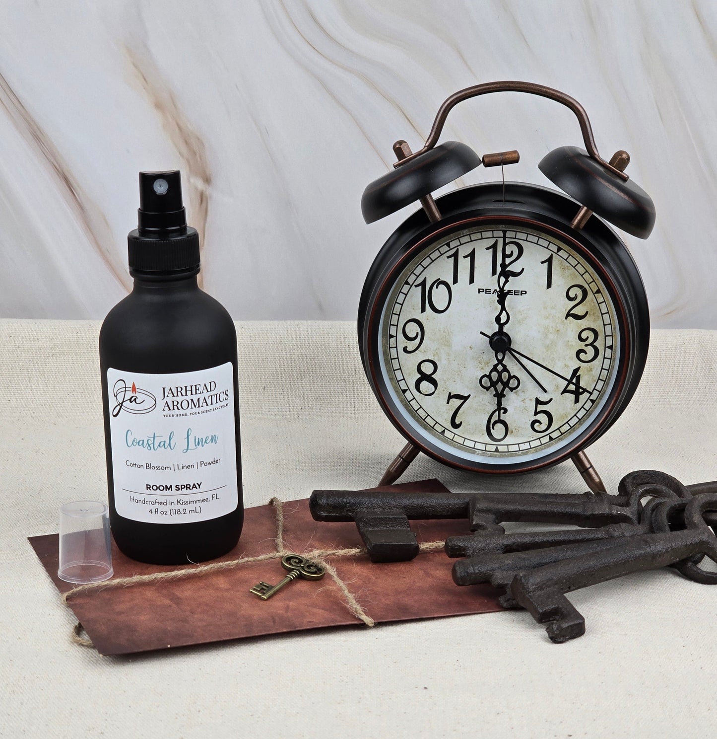 A vintage-style black alarm clock is next to Jarhead Aromatics Coastal Linen Room Spray on a brown surface, illustrating a coastal retreat. Nearby are old-fashioned keys and a small bronze key on leather. The light marbled background enhances the serene ambiance.