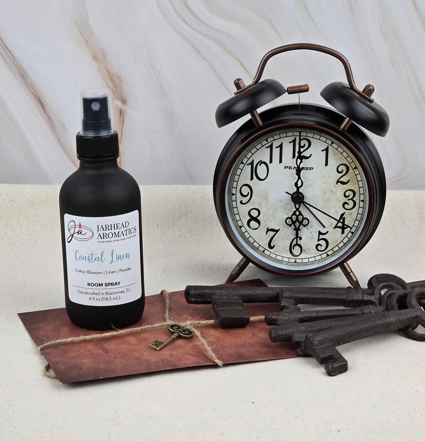 A vintage alarm clock sits on a table with a Coastal Linen - Room Spray by Jarhead Aromatics, a leather-bound notebook, and two large antique keys against a marble-patterned wall.