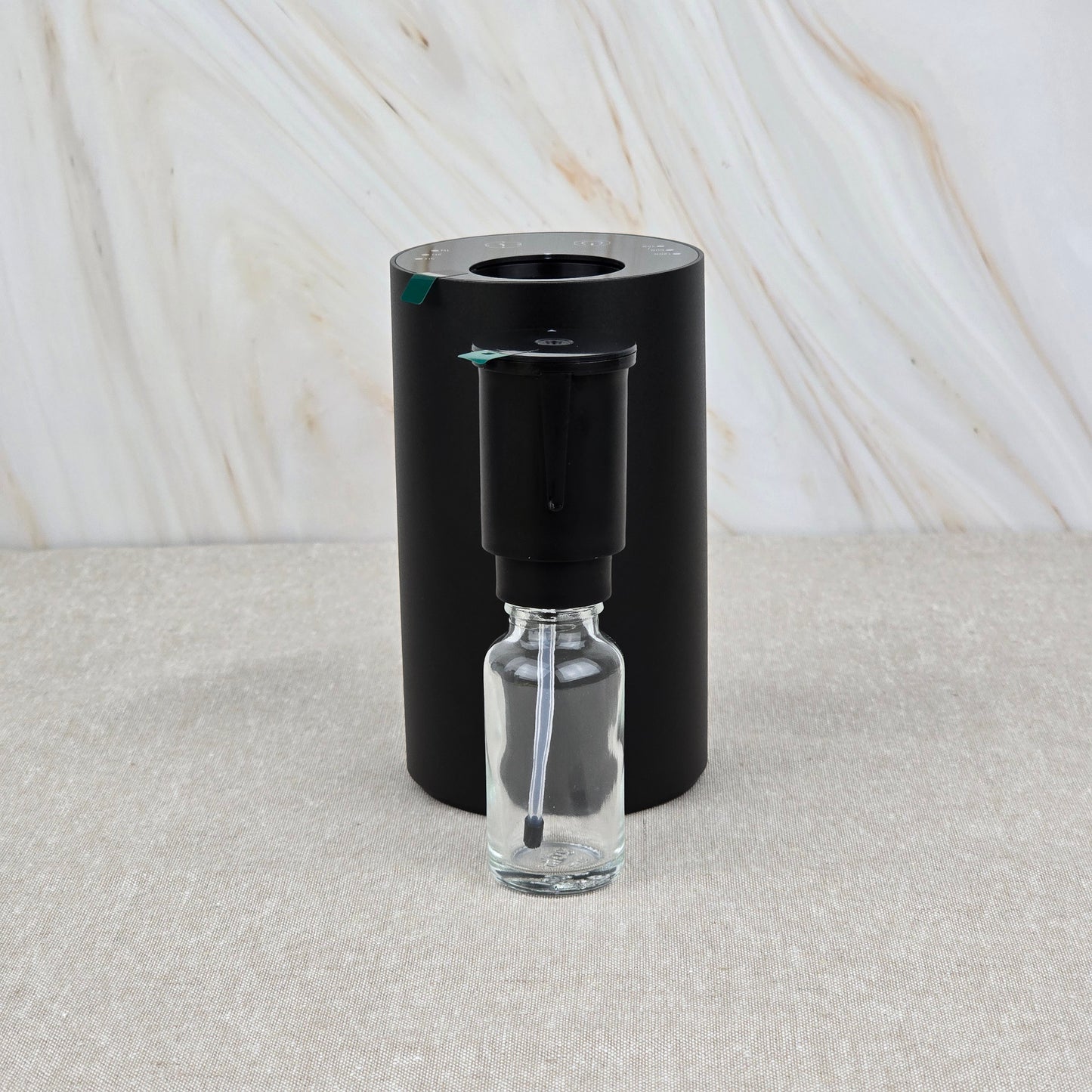 A small clear glass dropper bottle sits before the Jarhead Aromatics Cold Air Diffuser—Portable & Rechargeable, a black cylinder—set against a neutral marbled background.