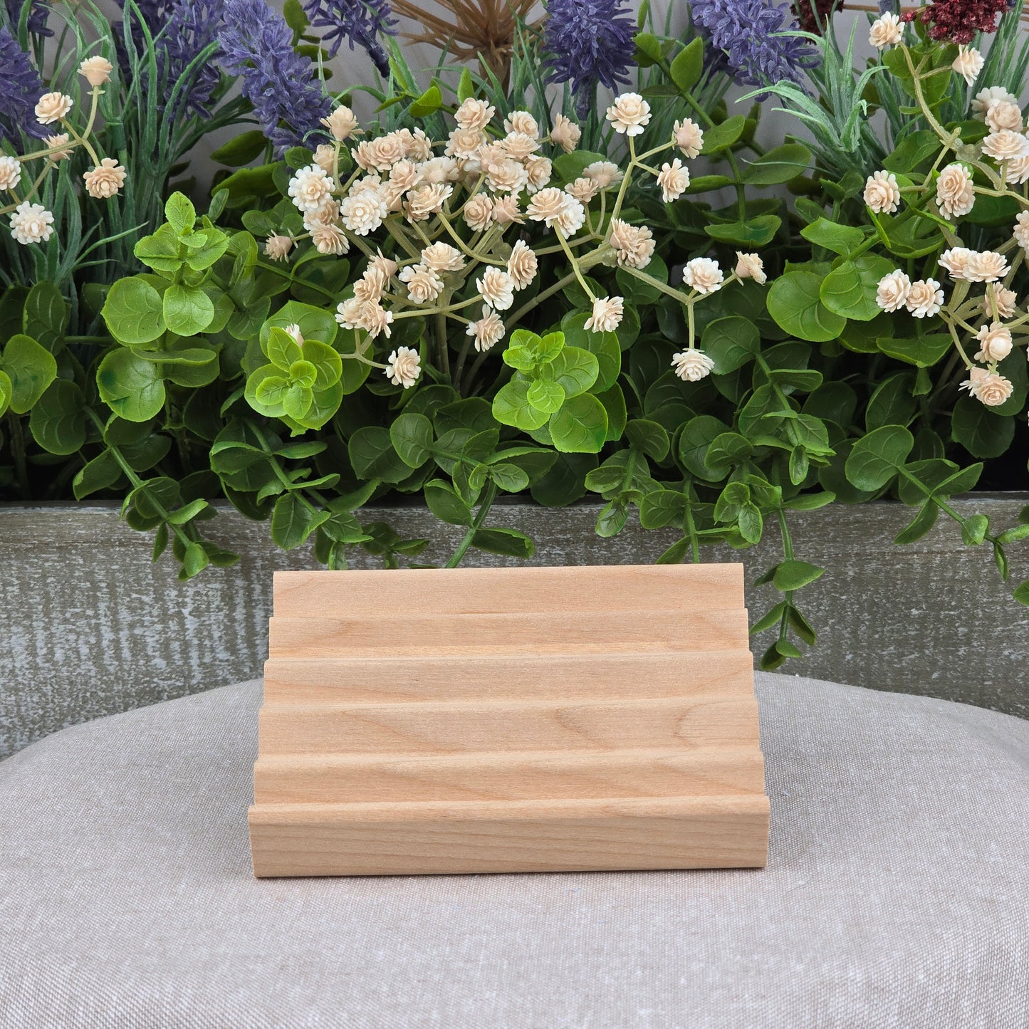 The Elevation Style Wood Soap Dish by Jarhead Aromatics, handcrafted from Red Alder with four grooves, rests on a gray cloth before faux green leaves and white-purple flowers—a sustainable, stylish soap saver for your bathroom.