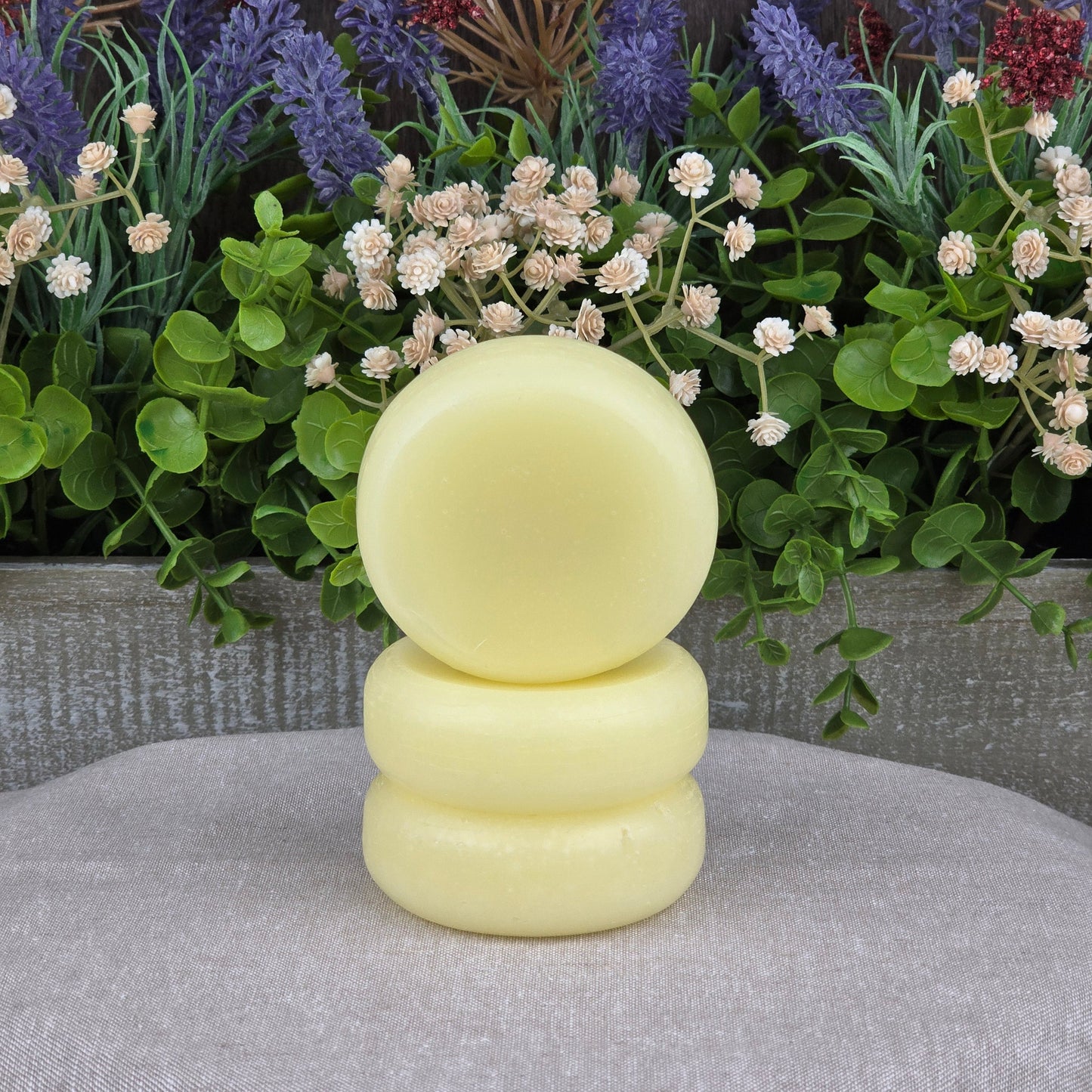 Three round, pale yellow Jarhead Aromatics Fragrance-Free Handmade Conditioner Bars are stacked before green leaves and colorful artificial flowers on a light gray surfaceโideal for anyone seeking zero-waste, all-natural hair care.
