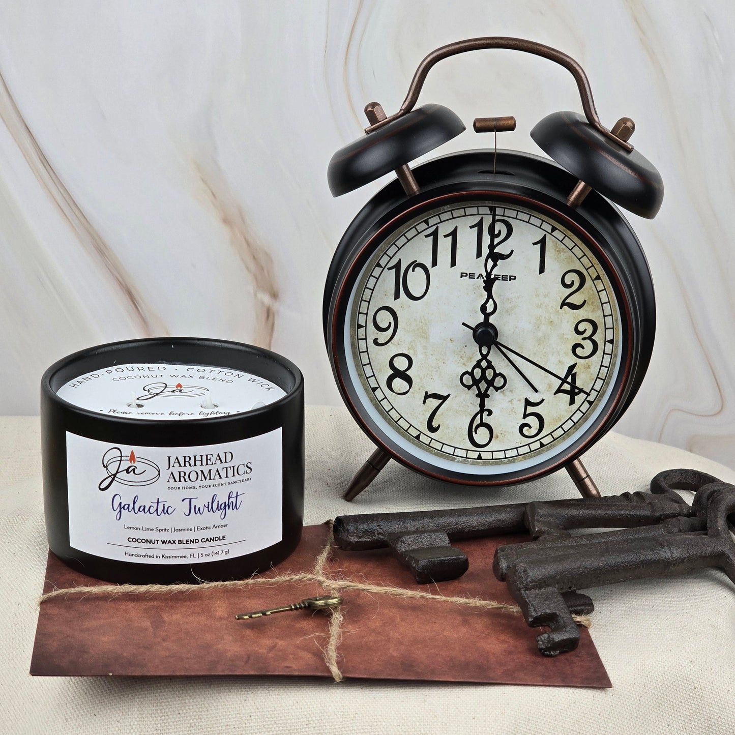 A black alarm clock reads 8:28 beside Jarhead Aromatics Galactic Twilight Coconut Wax Blend Candle from the Timeless Collection, a brown envelope topped with a gold key, and two large vintage keys on a marble-patterned light surface.