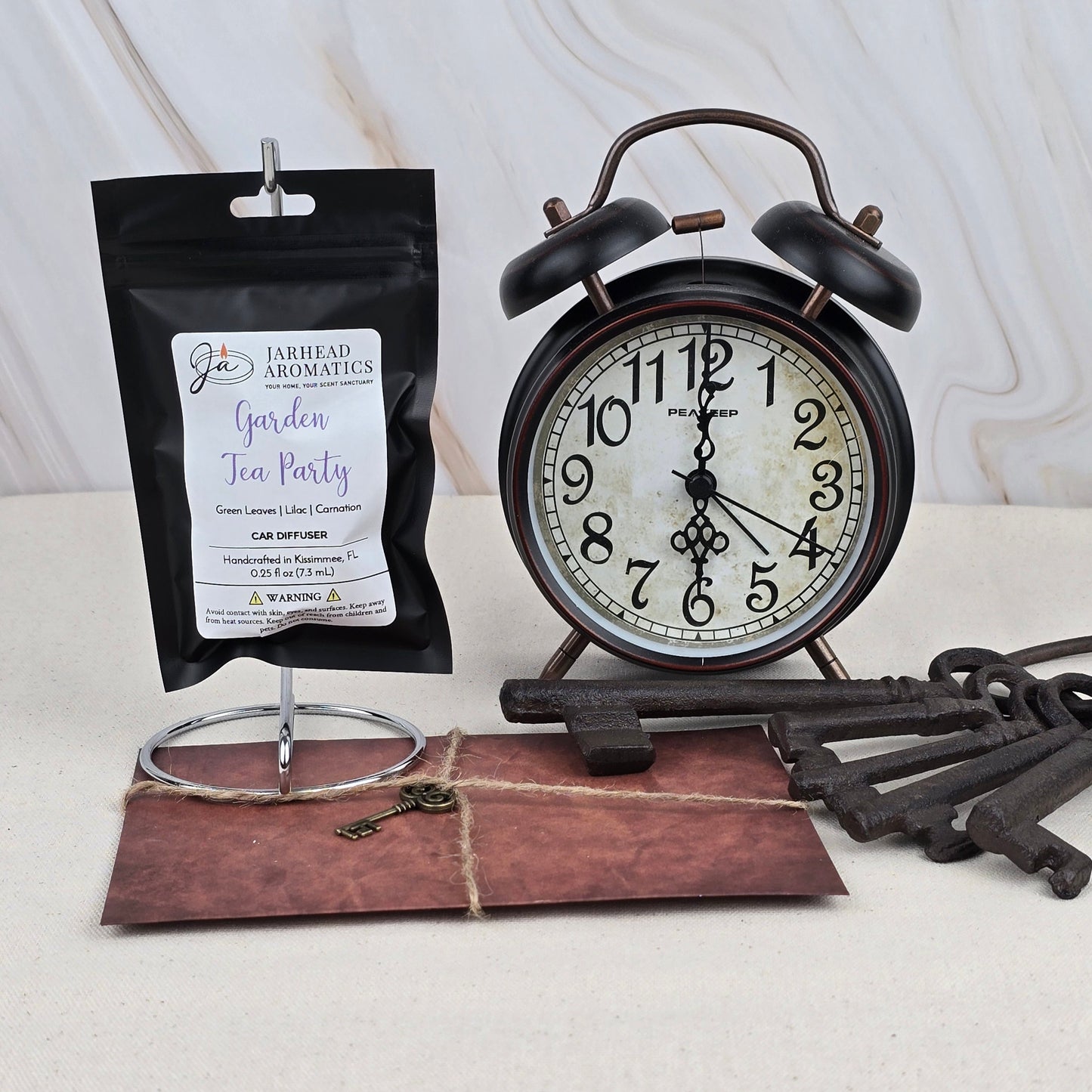 A Jarhead Aromatics Garden Tea Party Car Diffuser rests with vintage décor—black alarm clock, metal stand, old keys, and a brown envelope—on a textured surface, evoking the calm of fresh linen.