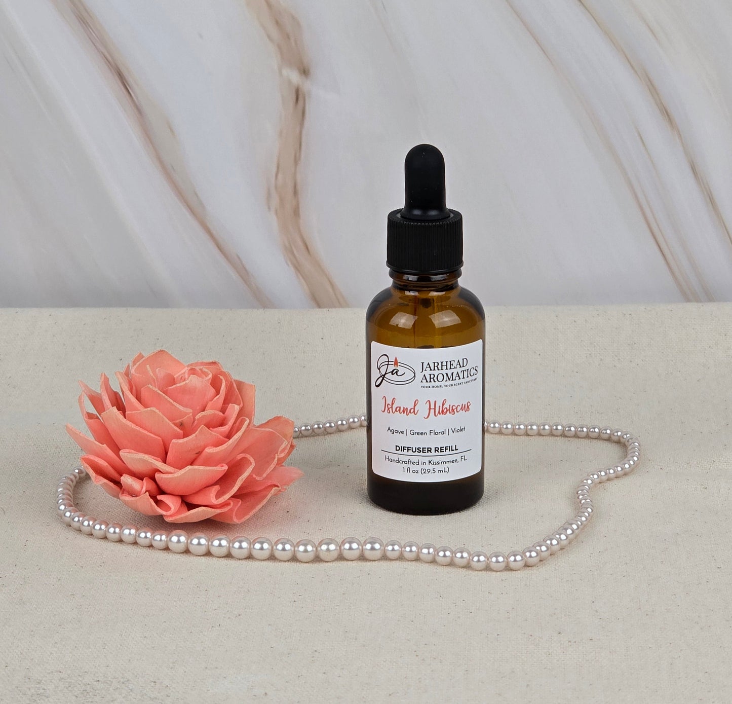 A small amber bottle, Island Hibiscus - Car Diffuser Refill by Jarhead Aromatics, with a dropper cap sits on a light surface exuding a tropical scent. Its surrounded by a pink flower and pearls against softly blurred marble-like textures in the background.