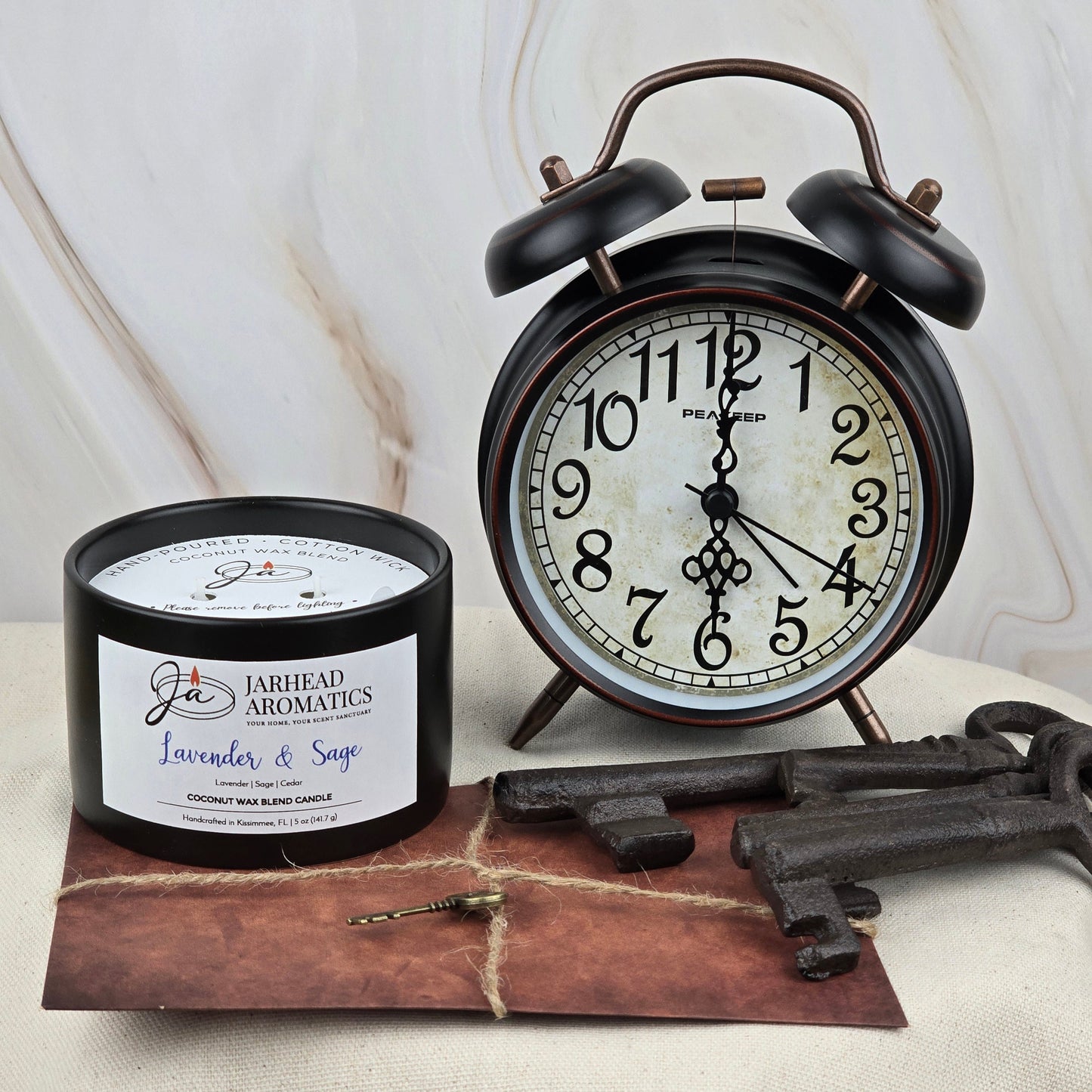 A black vintage alarm clock shows 6:00 beside the Jarhead Aromatics Timeless Collection Lavender & Sage Coconut Wax Blend Candle, evoking a subtle spa feel, with an old brown envelope and three antique keys nearby.