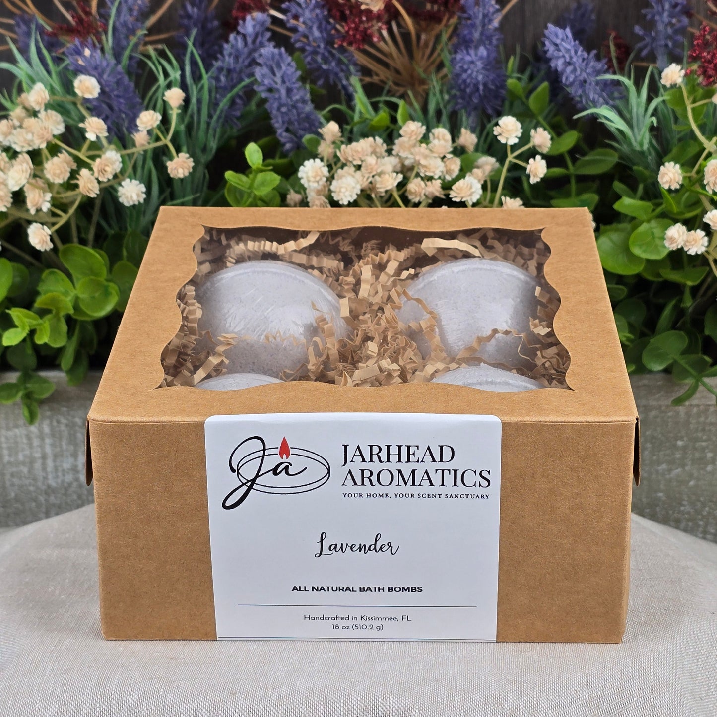A box of Jarhead Aromatics Patchouli Bath Bomb - All Natural sits on a gray surface, set against purple and white flowers. The brown box has a clear window displaying cruelty-free bath bombs and a white label on the front.
