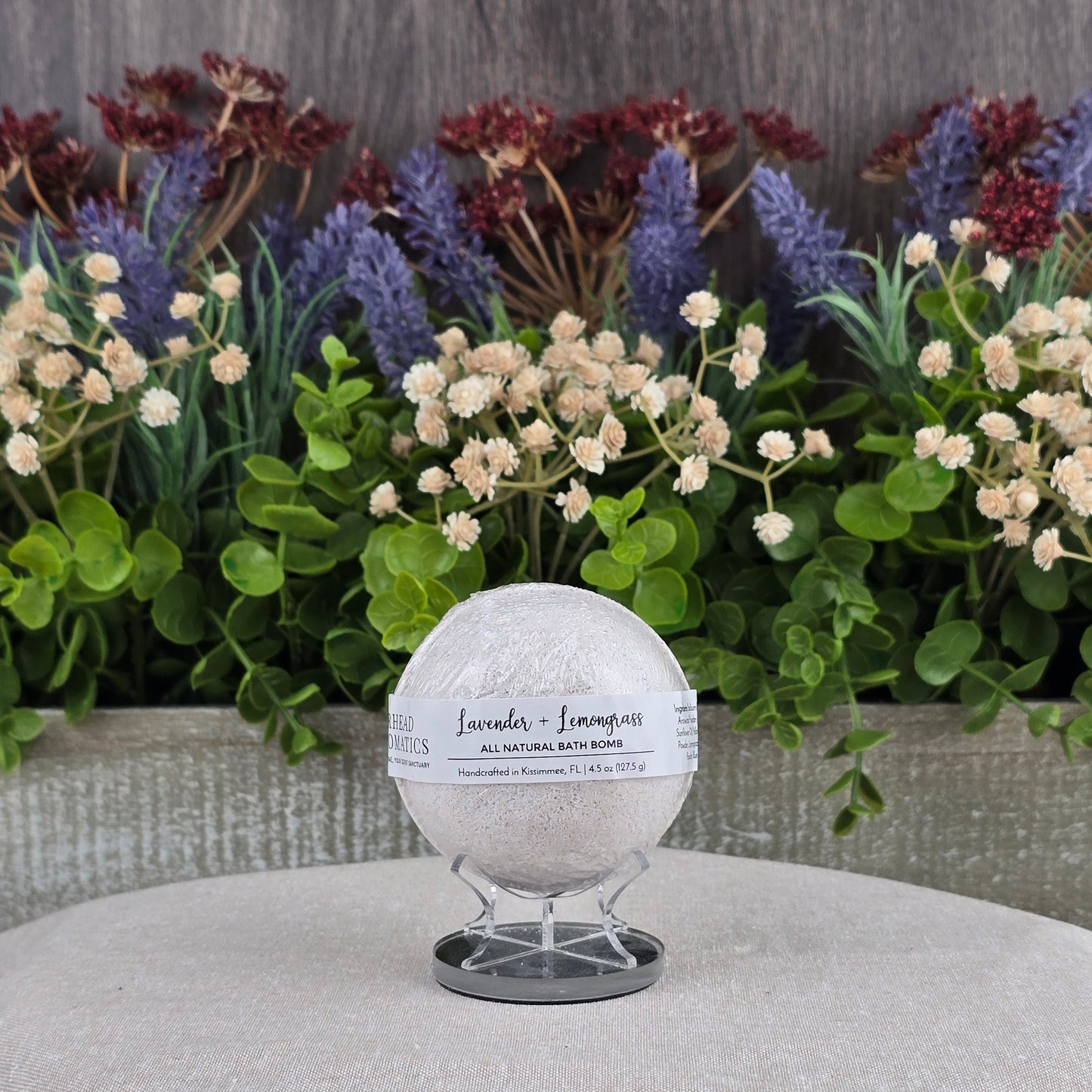 The Jarhead Aromatics Lavender + Lemongrass Bath Bomb - All Natural rests on a stand atop a beige cushion, with artificial flowers and greenery behind it to emphasize its natural ingredients.