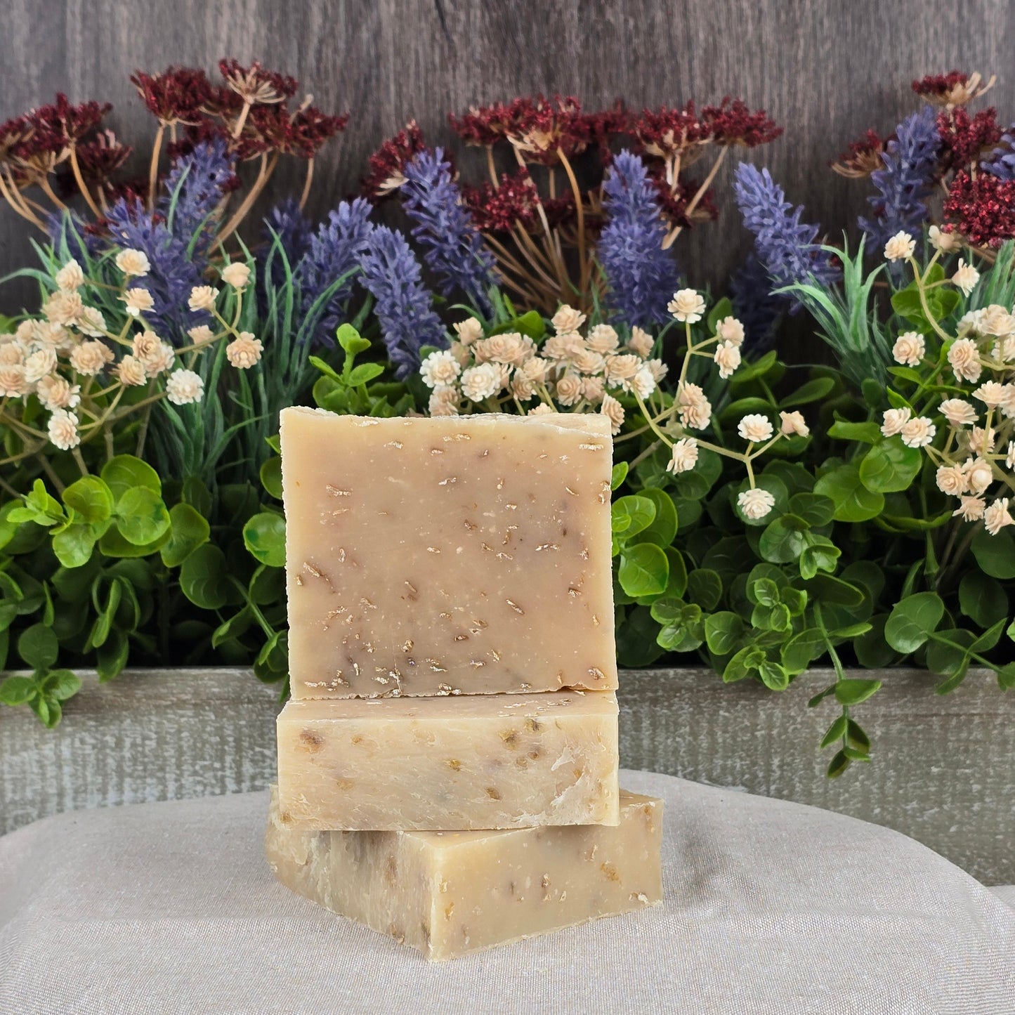 Three Lavender + Oatmeal Handmade Goat Milk Soap bars by Jarhead Aromatics are stacked against a backdrop of purple and red dried flowers with green leaves. Infused with natural skincare essences, the soap displays herb or oatmeal specks atop a light cloth.