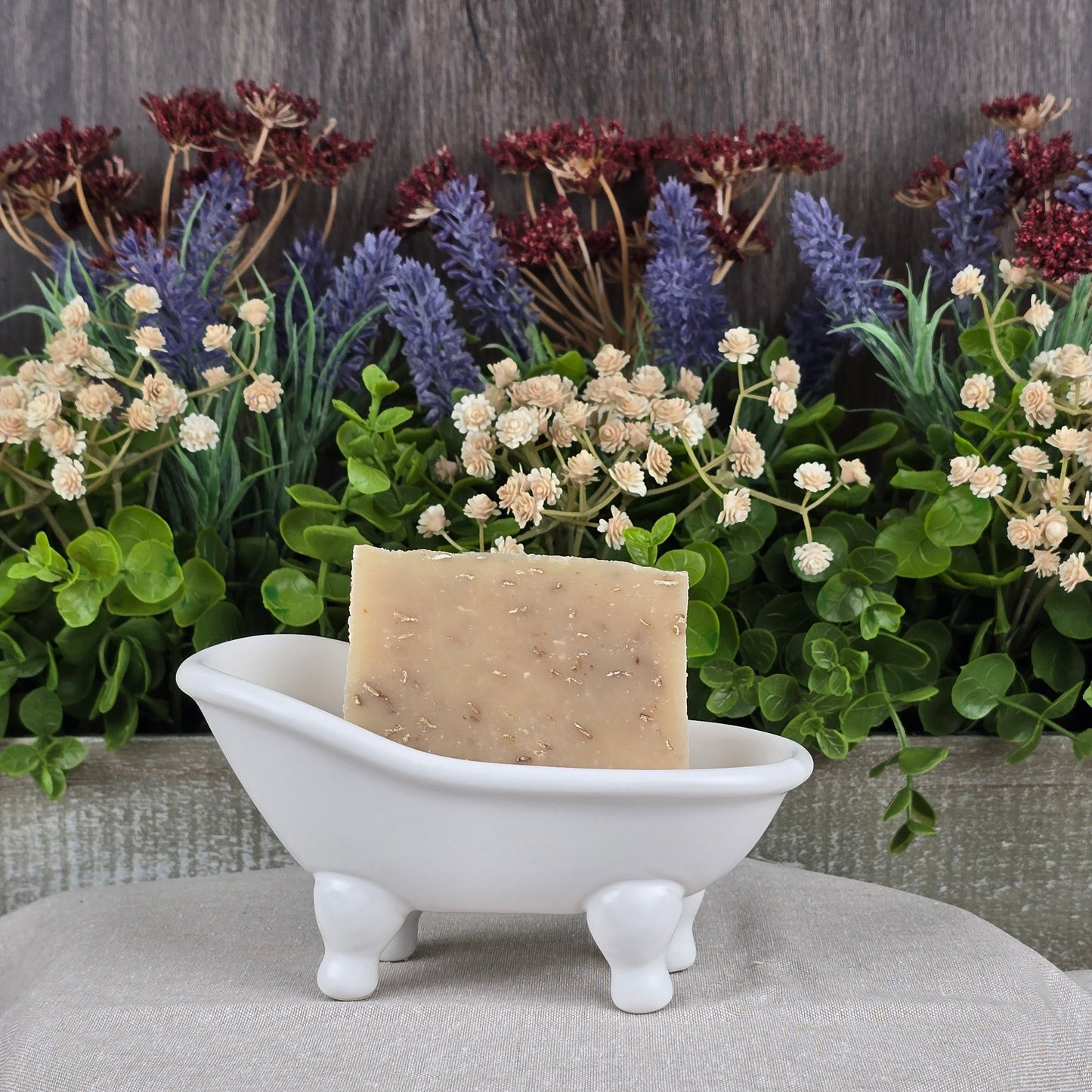 The Lavender + Oatmeal Handmade Goat Milk Soap by Jarhead Aromatics sits in a decorative white bathtub on a textured surface, complemented by vibrant artificial lavender and red-tipped foliage, adding rustic charm against a wooden backdrop.