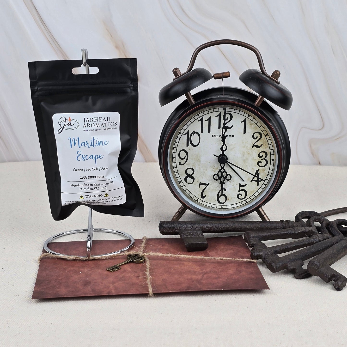 A black alarm clock, a Jarhead Aromatics Maritime Escape Car Diffuser packet, an old envelope with a key on top, and large vintage-style keys are arranged on a light surface against a marbled background, evoking fresh citrus scents.