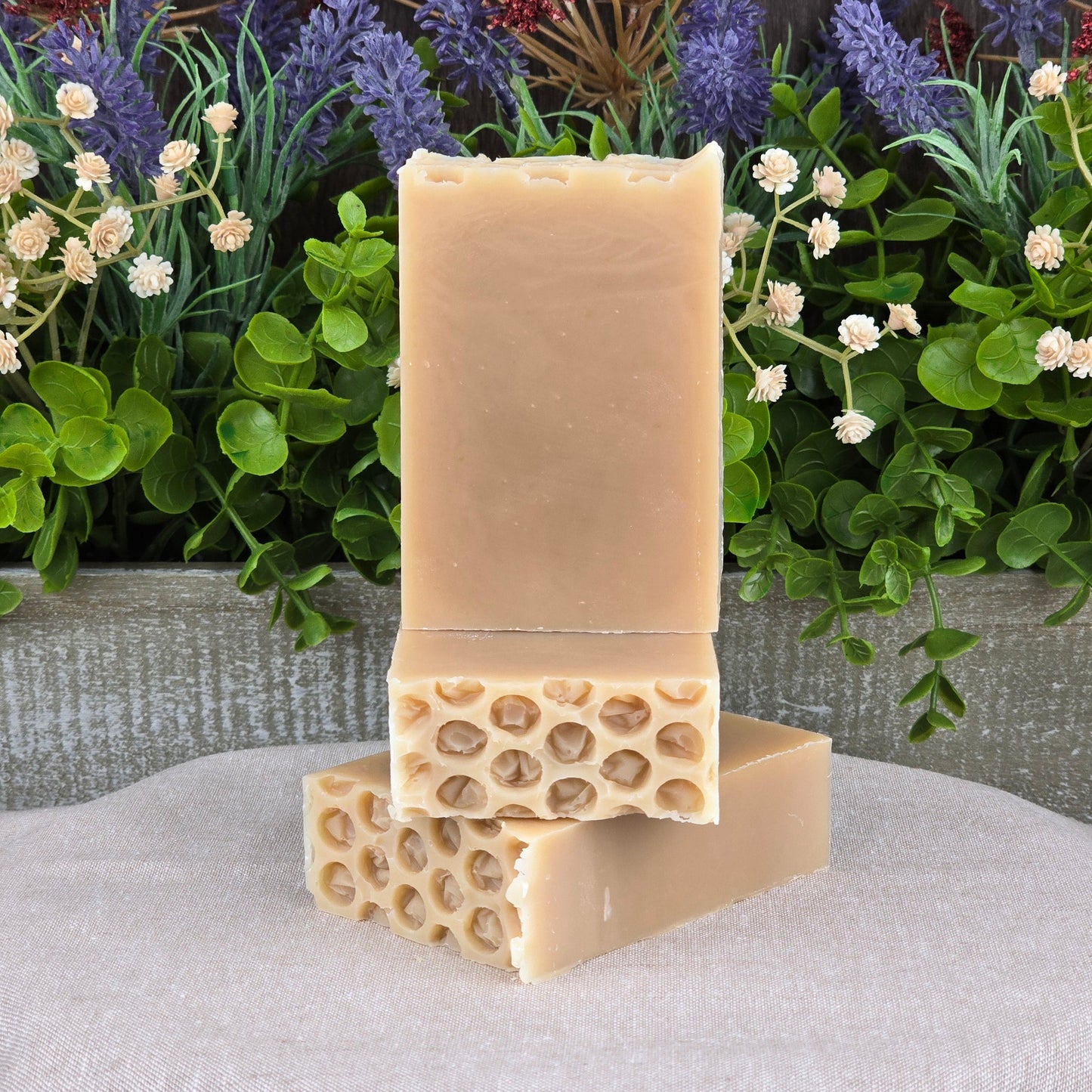 The Milk + Honey Handmade Artisan Soap by Jarhead Aromatics, a rectangular beige natural bar, stands upright on two similar honeycomb-patterned bars, with green leaves, white flowers, and purple lavender on a neutral cloth in the background.