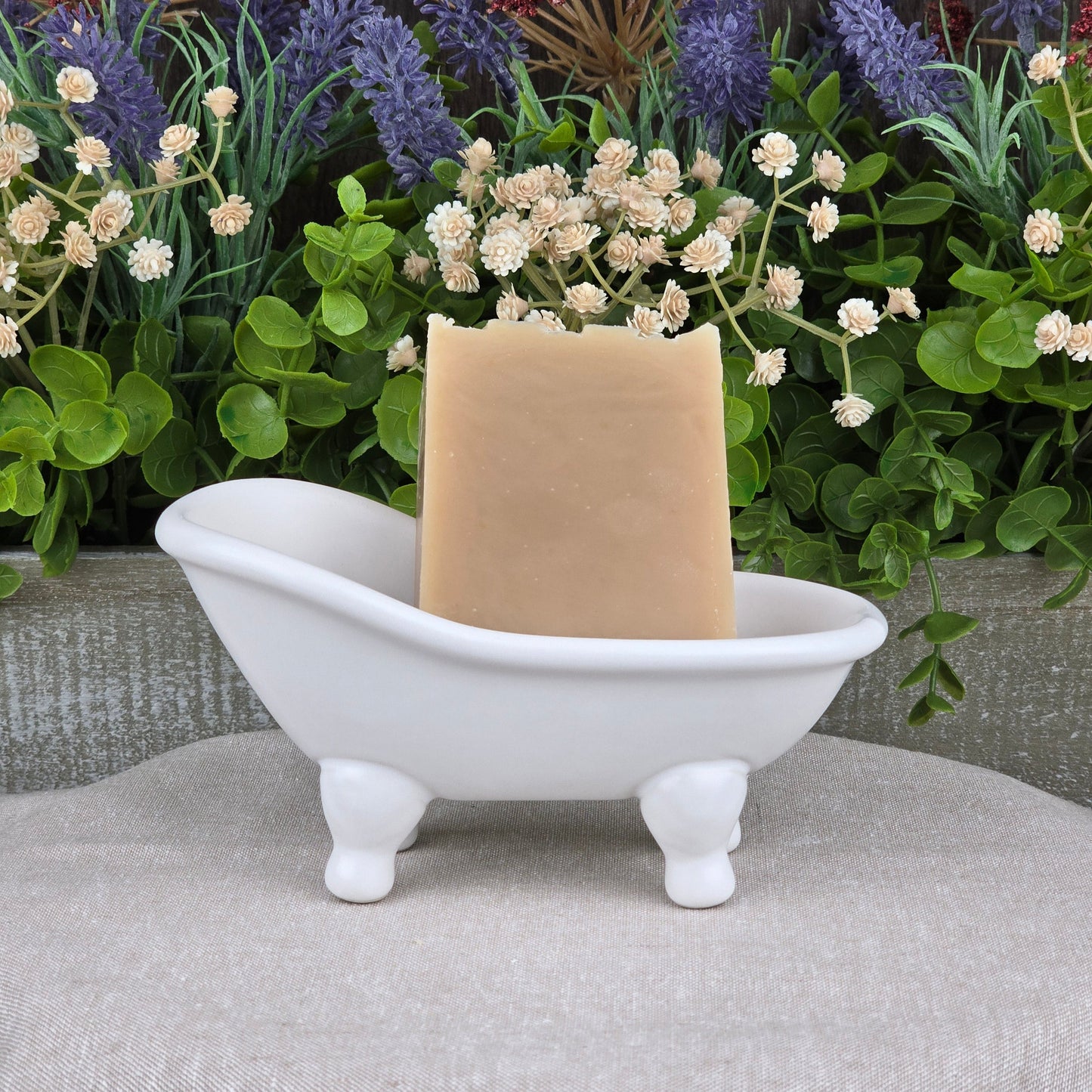 The Milk + Honey Handmade Artisan Soap by Jarhead Aromatics sits upright in a small white clawfoot bathtub soap dish, with artificial green plants and white flowers in the background.