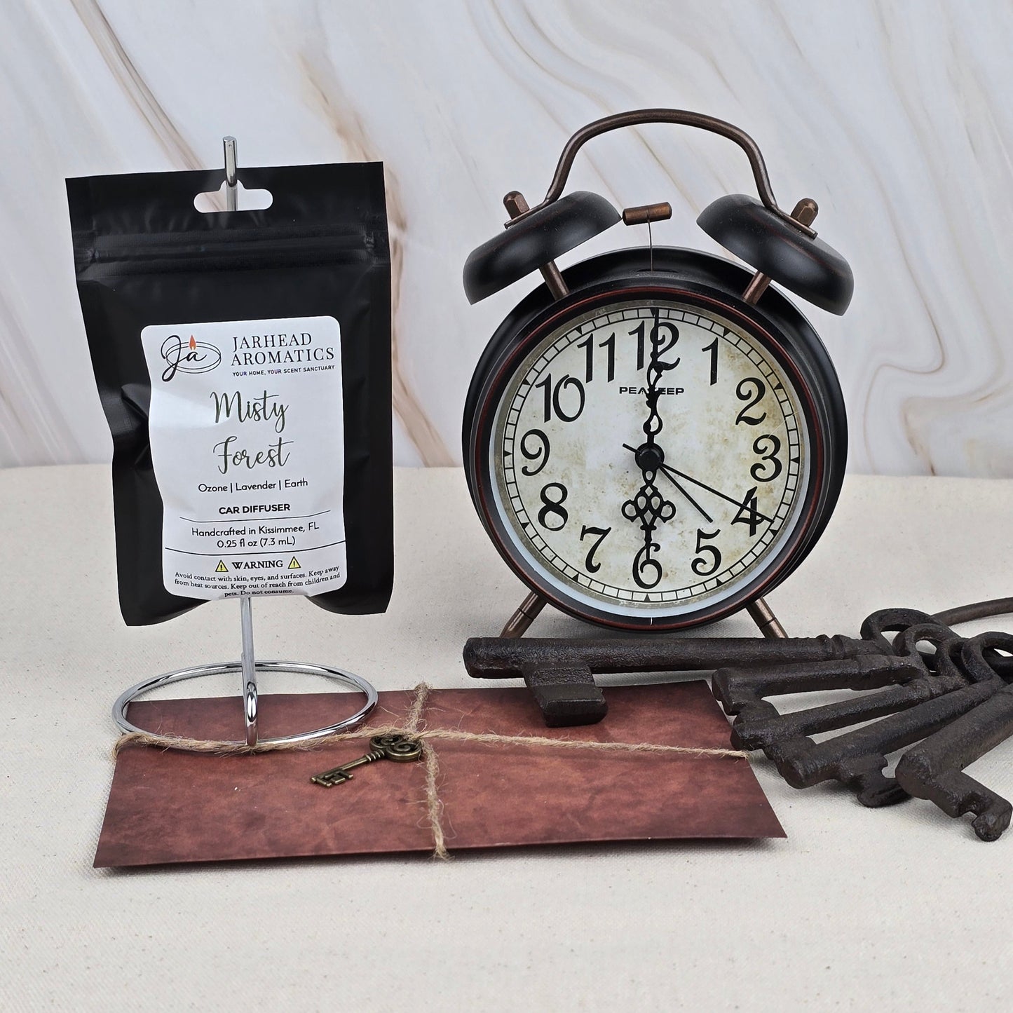 A vintage black alarm clock, a bundle of old metal keys, and the Misty Forest Car Diffuser by Jarhead Aromatics rest on a light surface against a marbled background.