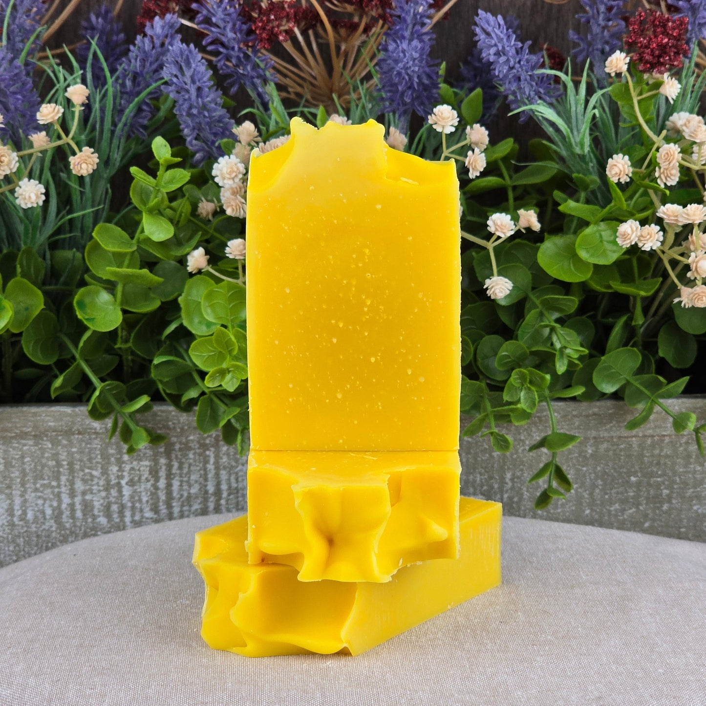 The Jarhead Aromatics Orange + Carrot Handmade Artisan Soapโan all-natural bar with wavy decorative edgesโis stacked atop two more artisan soaps, set against lush green leaves and delicate white and purple flowers.