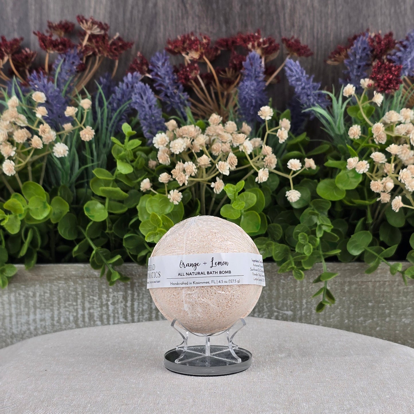 The Orange + Lemon Bath Bomb - All Natural by Jarhead Aromatics sits on a stand atop a gray cushion, with artificial green leaves and colorful flowers behind it, highlighting its essential oils and natural ingredients.