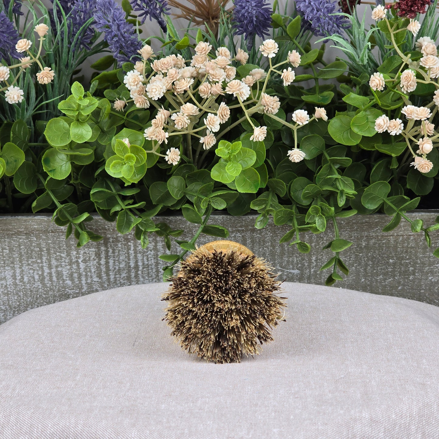 The Jarhead Aromatics Bamboo Handle Palm Fiber Palm Scrubber Brush stands upright on a light gray fabric, with green leaves and white and purple artificial flowers in the background.
