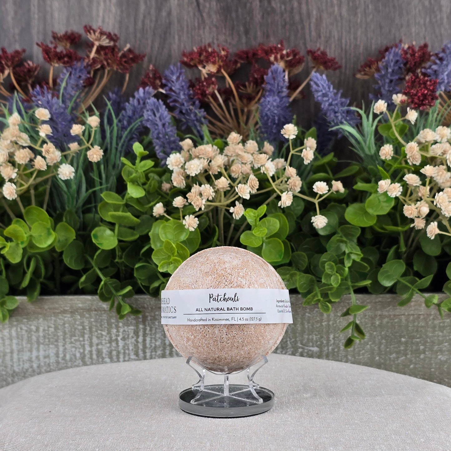 The Patchouli Bath Bomb - All Natural by Jarhead Aromatics rests on a clear stand, surrounded by green leaves and purple and cream flowers atop a gray surface. Made with natural ingredients for a soothing bath experience.