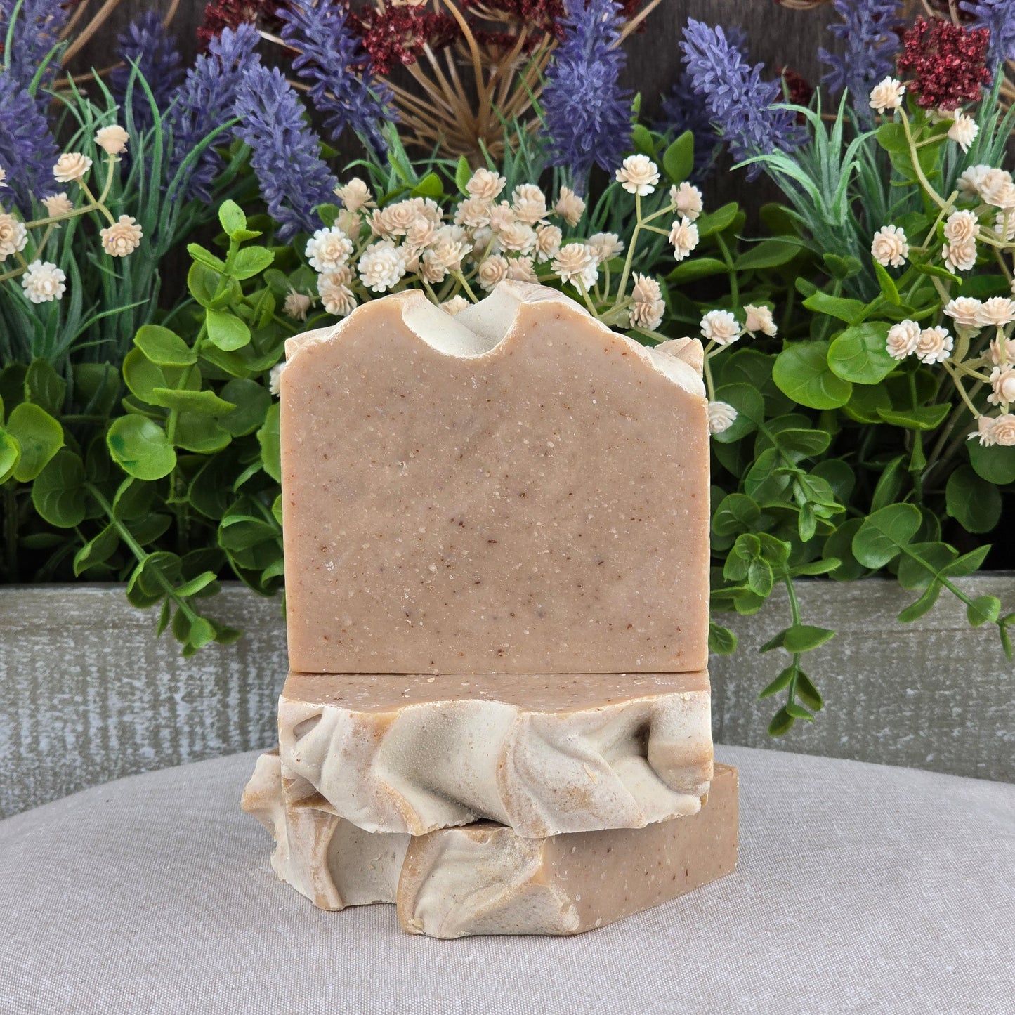 Two bars of Jarhead Aromaticsโ Pumpkin + Spice Handmade Seasonal Artisan Soap, one upright and one flat, are shown with lush greenery and purple flowers. The cold process soap has a smooth finish and a wavy, decorative edge.