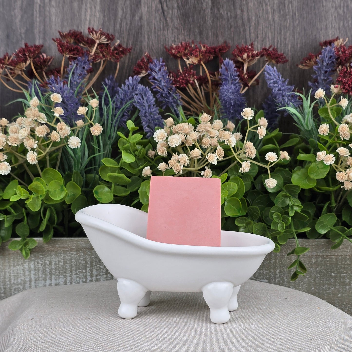 A small white, clawfoot bathtub-shaped dish holds a pink Rose + Calamine Handmade Goat Milk Facial Soap by Jarhead Aromatics. Behind it is a display of assorted artificial flowers and greenery against a wooden background on a textured cloth surface, highlighting the elegance of nourishing oils.