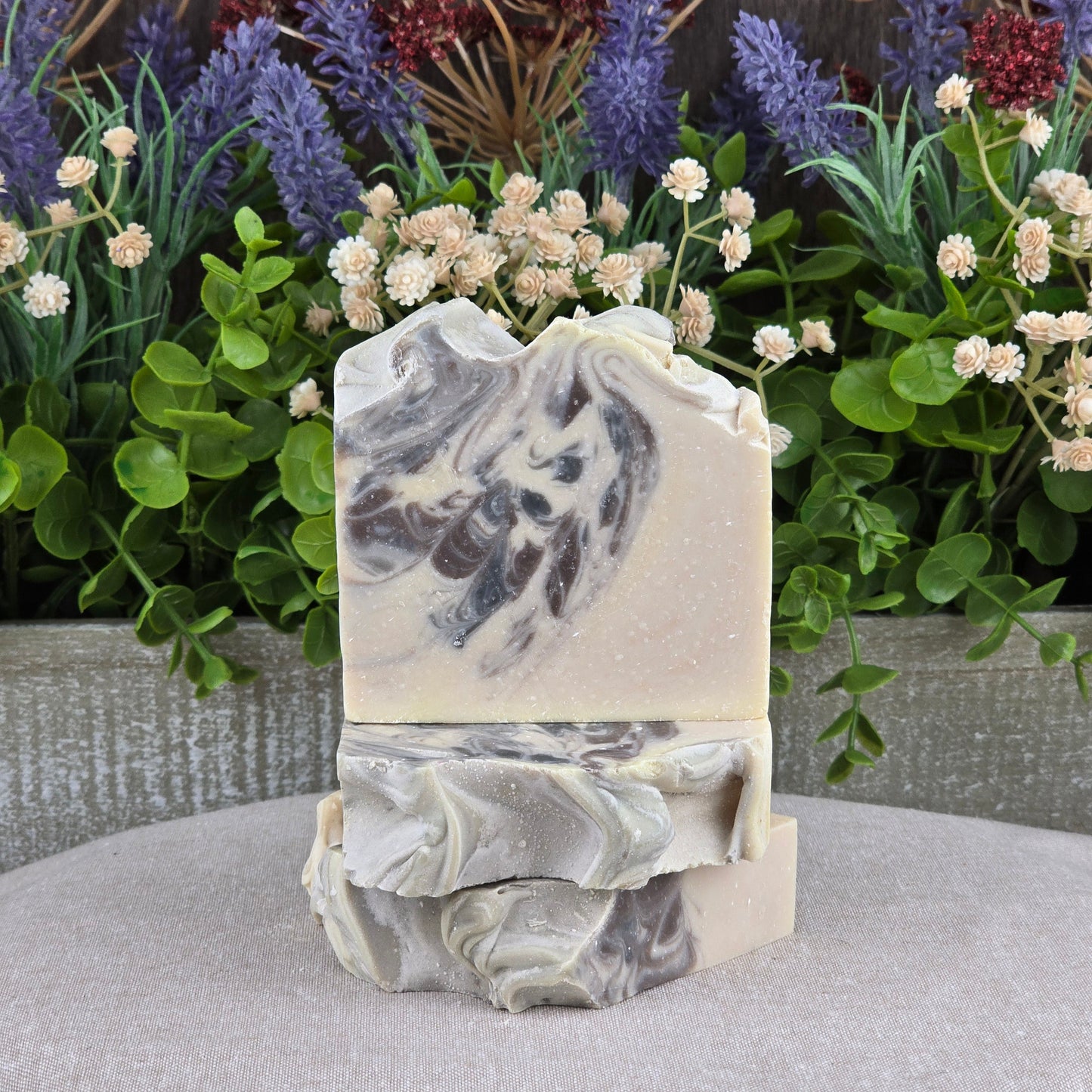 Two bars of Jarhead Aromatics Sandalwood + Patchouli Handmade Artisan Soap, with beige and dark brown swirls, are stacked in front of green and purple artificial flowers on a light fabric surface.