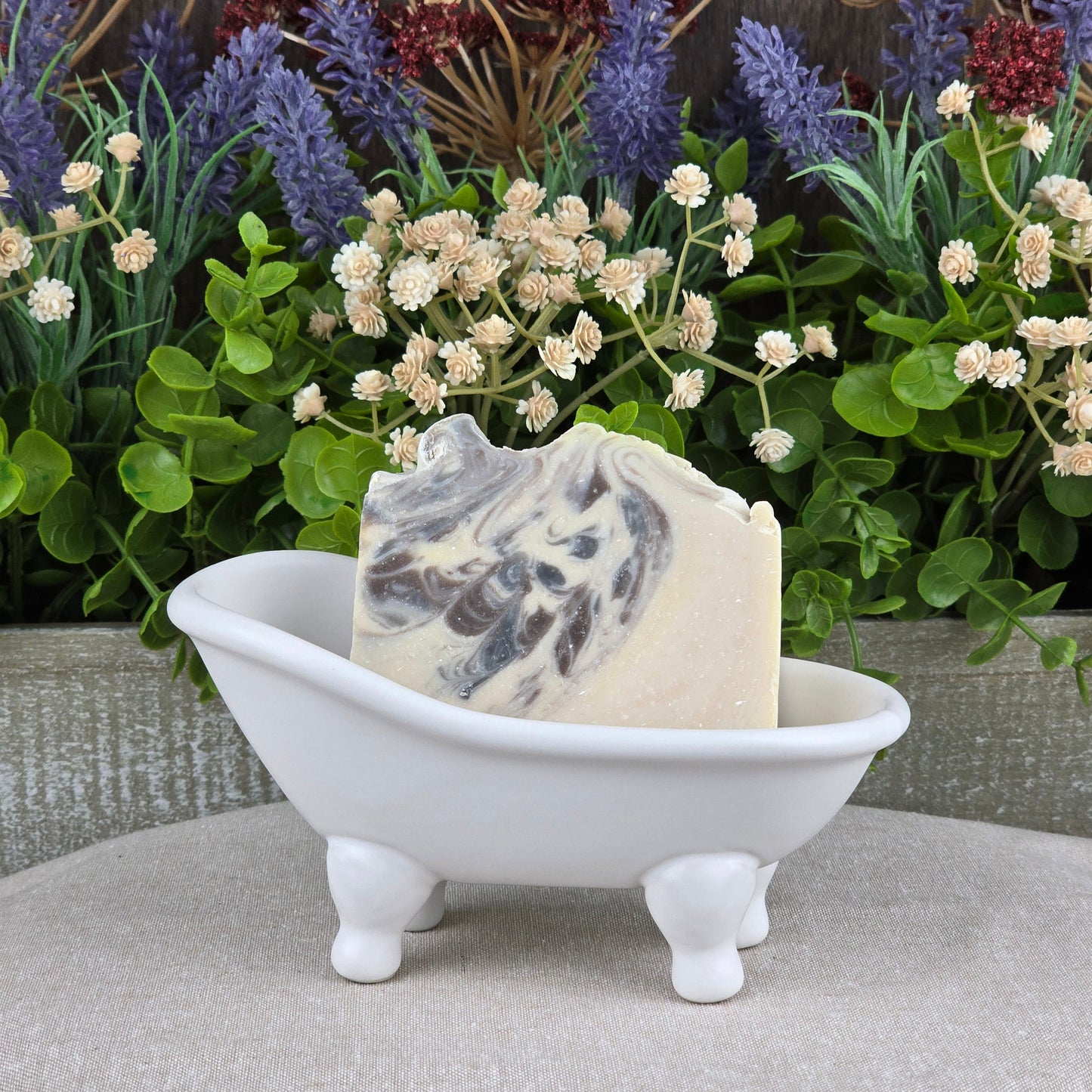 The Jarhead Aromatics Sandalwood + Patchouli Handmade Artisan Soap rests in a small white clawfoot bathtub dish, set before lush green plants and purple flowers.