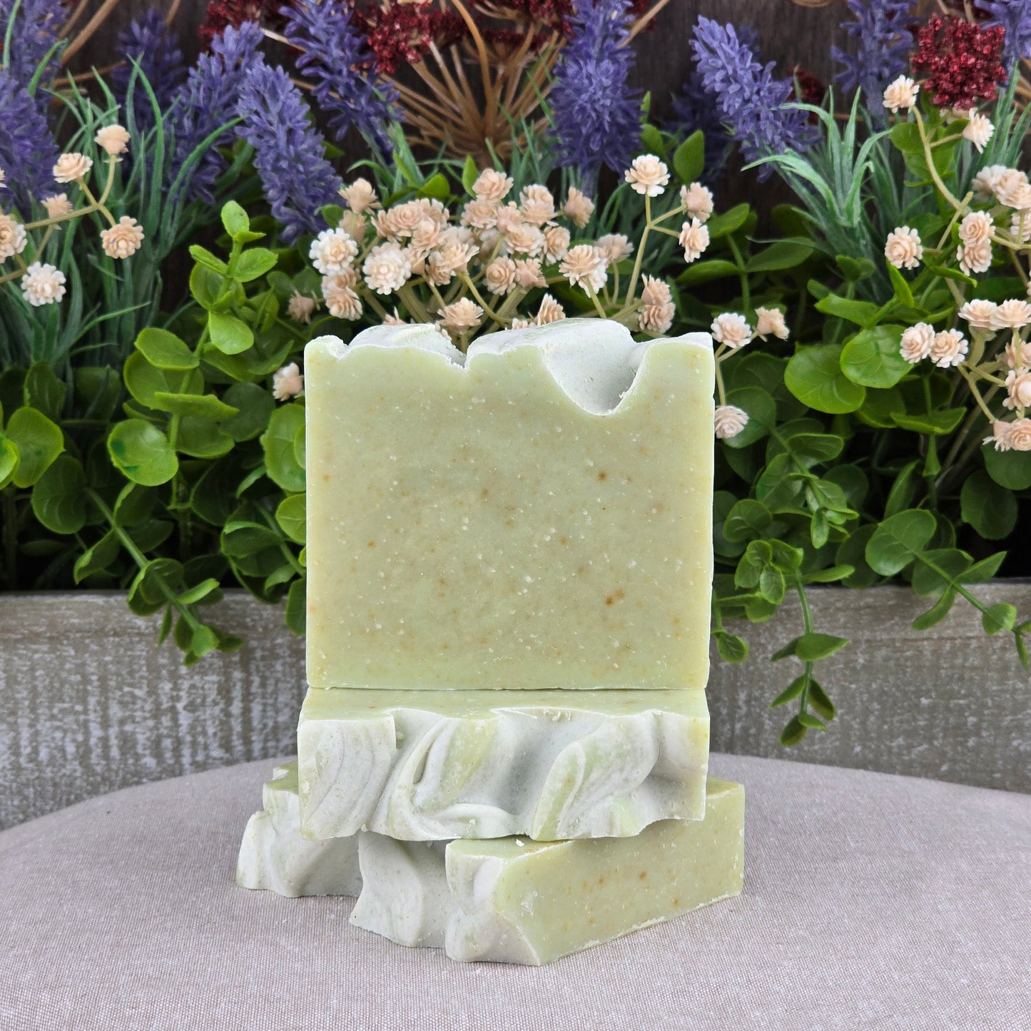 Three bars of Jarhead Aromatics Sea Kelp + Aloe Handmade Artisan Soap are stacked before green leaves and purple-white flowers, highlighting the soapโs all-natural, artisan quality in a lush, rustic setting.