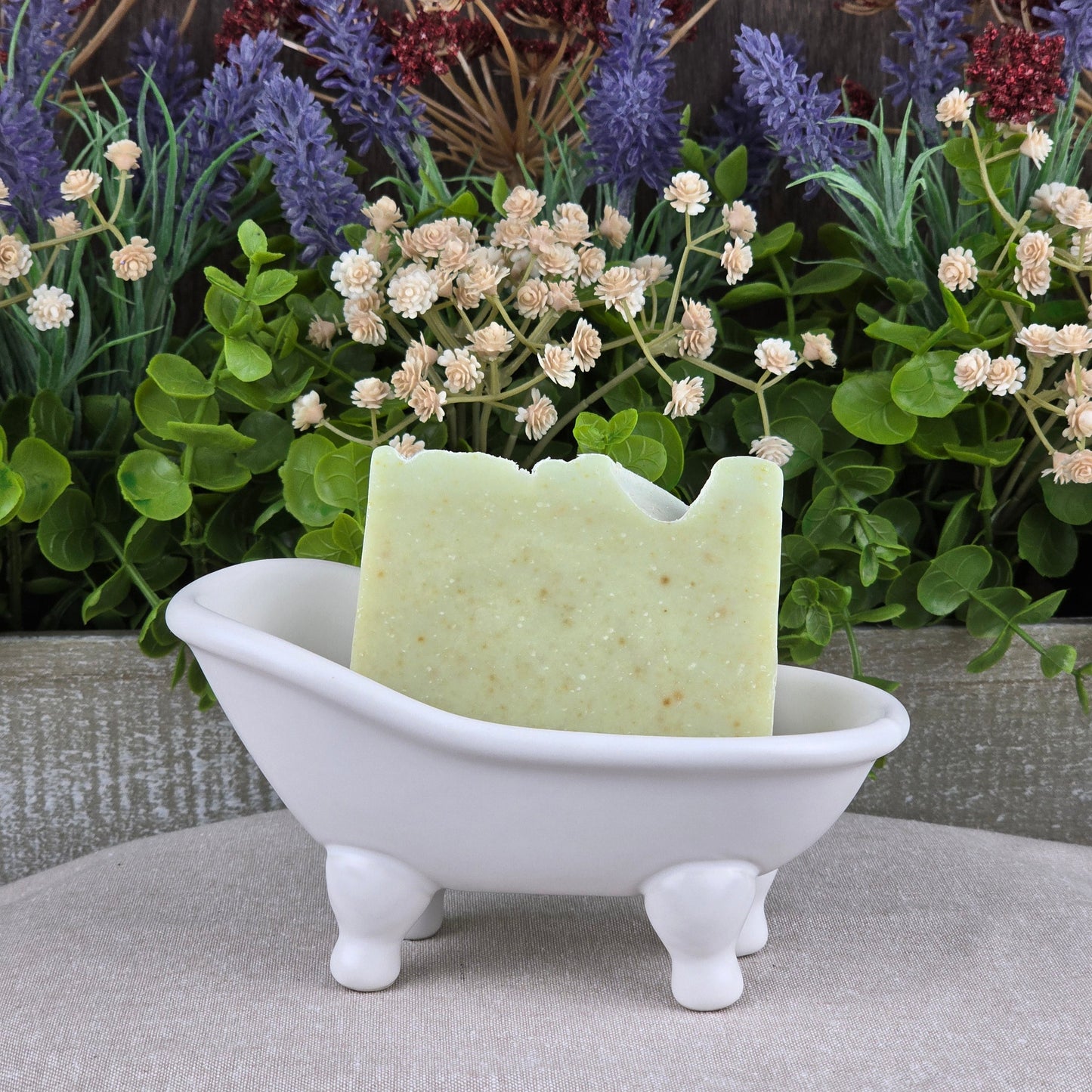 A bar of Jarhead Aromatics' Sea Kelp + Aloe Handmade Artisan Soap sits in a small white bathtub-shaped dish, with artificial greenery and purple and white flowers in the background.