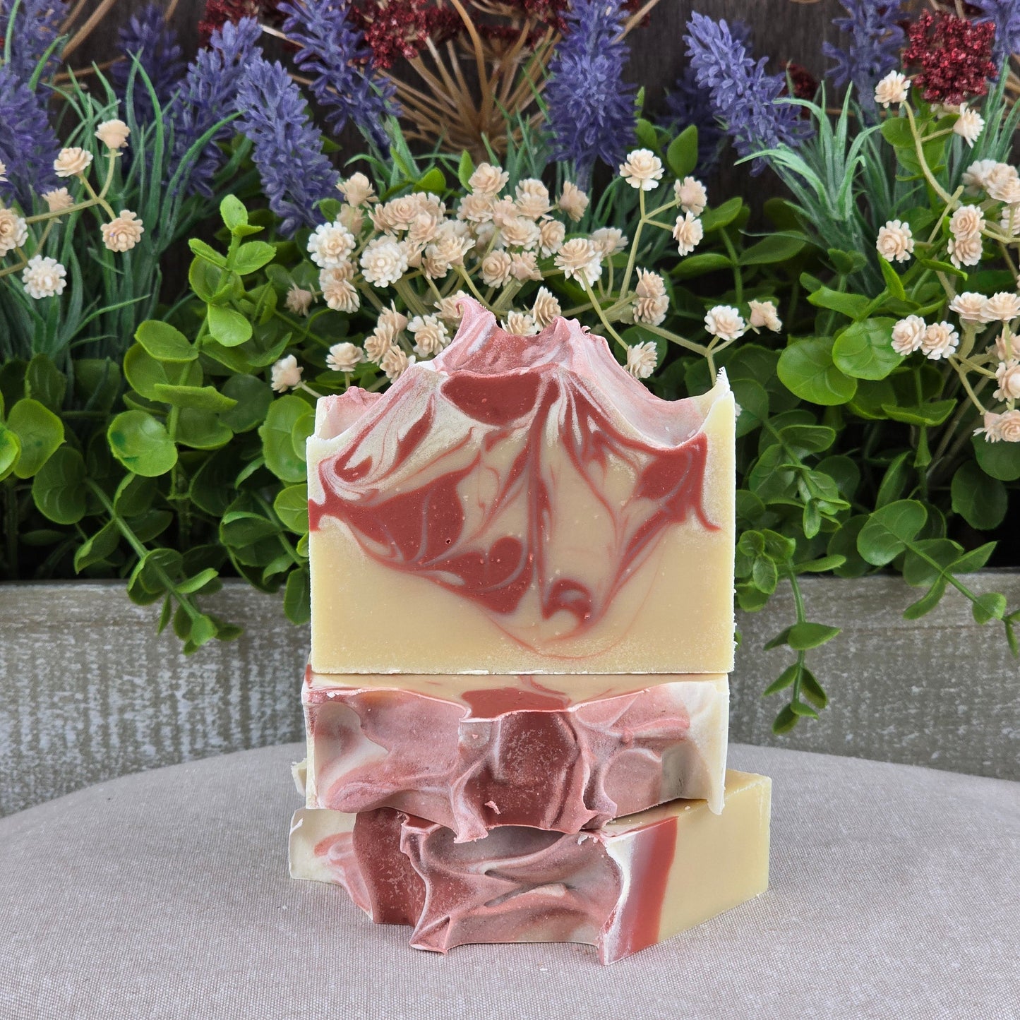 Three Sea Moss + Aloe Handmade Artisan Soaps from Jarhead Aromatics, with cream and mauve swirls, are stacked before green leaves, lavender, and white flowersโideal for anyone who appreciates all-natural artisan blends.