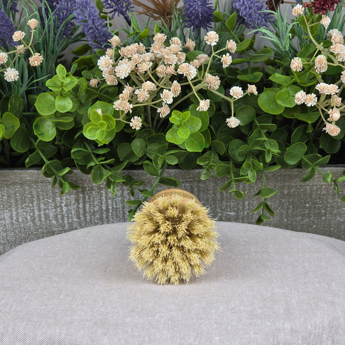 The Jarhead Aromatics Bamboo Handle Sisal Fiber Palm Scrubber Brush, a natural kitchen dish and produce cleaning brush, is displayed on a beige fabric with green leaves and white and purple flowers in the background.