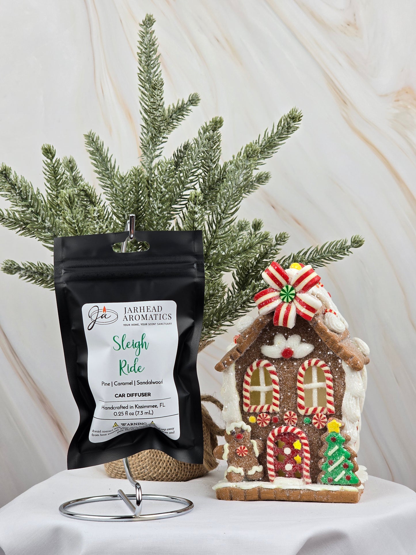 A Jarhead Aromatics Sleigh Ride car diffuser is on a metal stand next to a gingerbread house with colorful candies. In the background, a frosted evergreen tree enhances the festive scene, capturing winters enchanting scent.