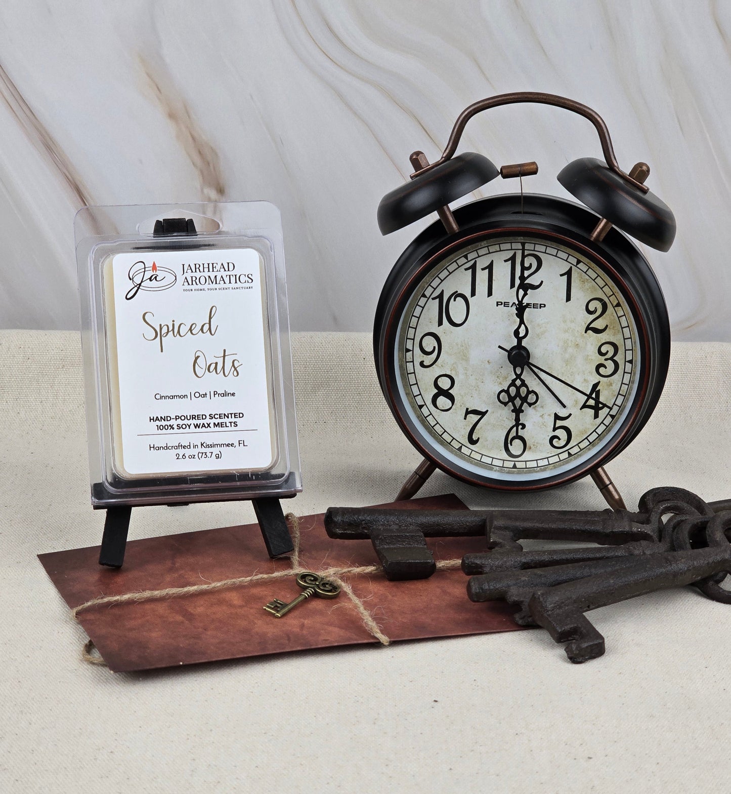 A vintage black alarm clock sits by a pack of Jarhead Aromatics Spiced Oats - Soy Wax Melt exuding a rich gourmand fragrance on a small easel. An old set of iron keys rests on a leather pouch against a light-colored, textured background.