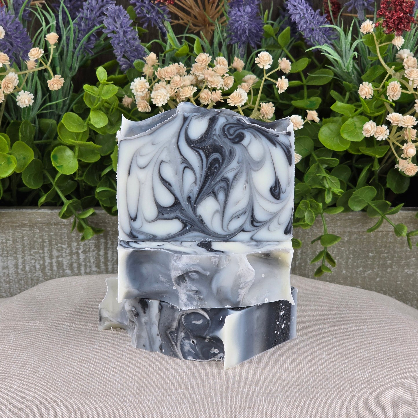 Jarhead Aromaticsโ Tea Tree + Tallow Handmade Artisan Soap bars, featuring detailed black, white, and gray marbling, are stacked on beige fabric with green plants and purple-cream flowers in the background for a natural look.