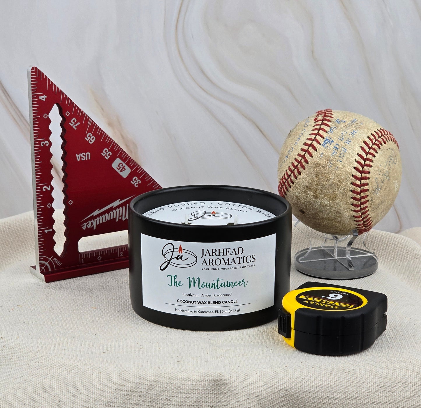 The Mountaineer - Coconut Wax Blend Candle by Jarhead Aromatics sits on fabric, surrounded by a red triangular measure, a displayed baseball, and a yellow/black tape measure, exuding the rustic cedarwood essence in its warm embrace.