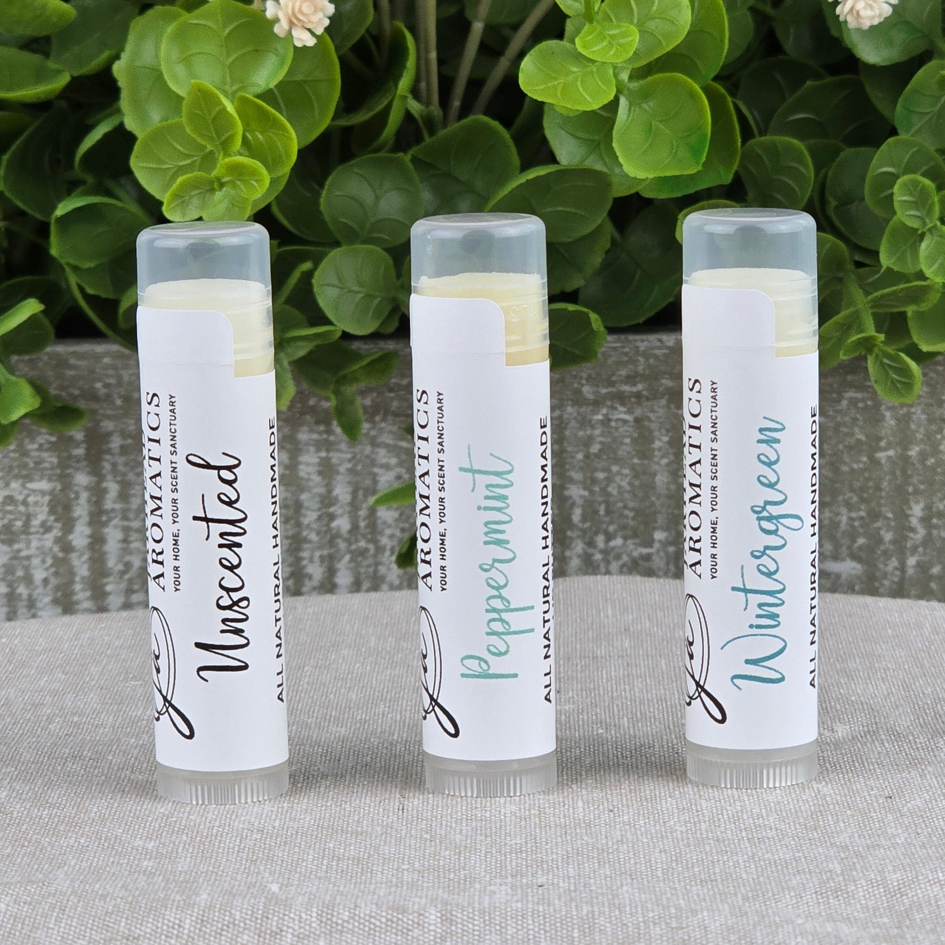 Three upright tubes of Jarhead Aromatics’ Lip Butter - All Natural in Unscented, Peppermint, and Wintergreen sit on gray cloth, surrounded by green leaves and cream flowers, highlighting their ultra-nourishing natural formula.
