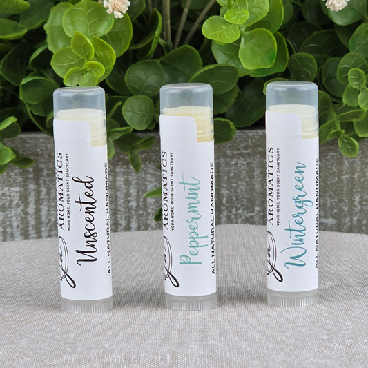 Three upright tubes of Jarhead Aromatics’ Lip Butter - All Natural in Unscented, Peppermint, and Wintergreen sit on gray cloth, surrounded by green leaves and cream flowers, highlighting their ultra-nourishing natural formula.