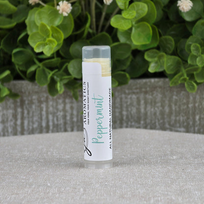 A tube of Jarhead Aromatics Lip Butter - All Natural stands upright on a gray fabric surface, with green leaves and small white flowers blurred in the background, suggesting its natural origins.