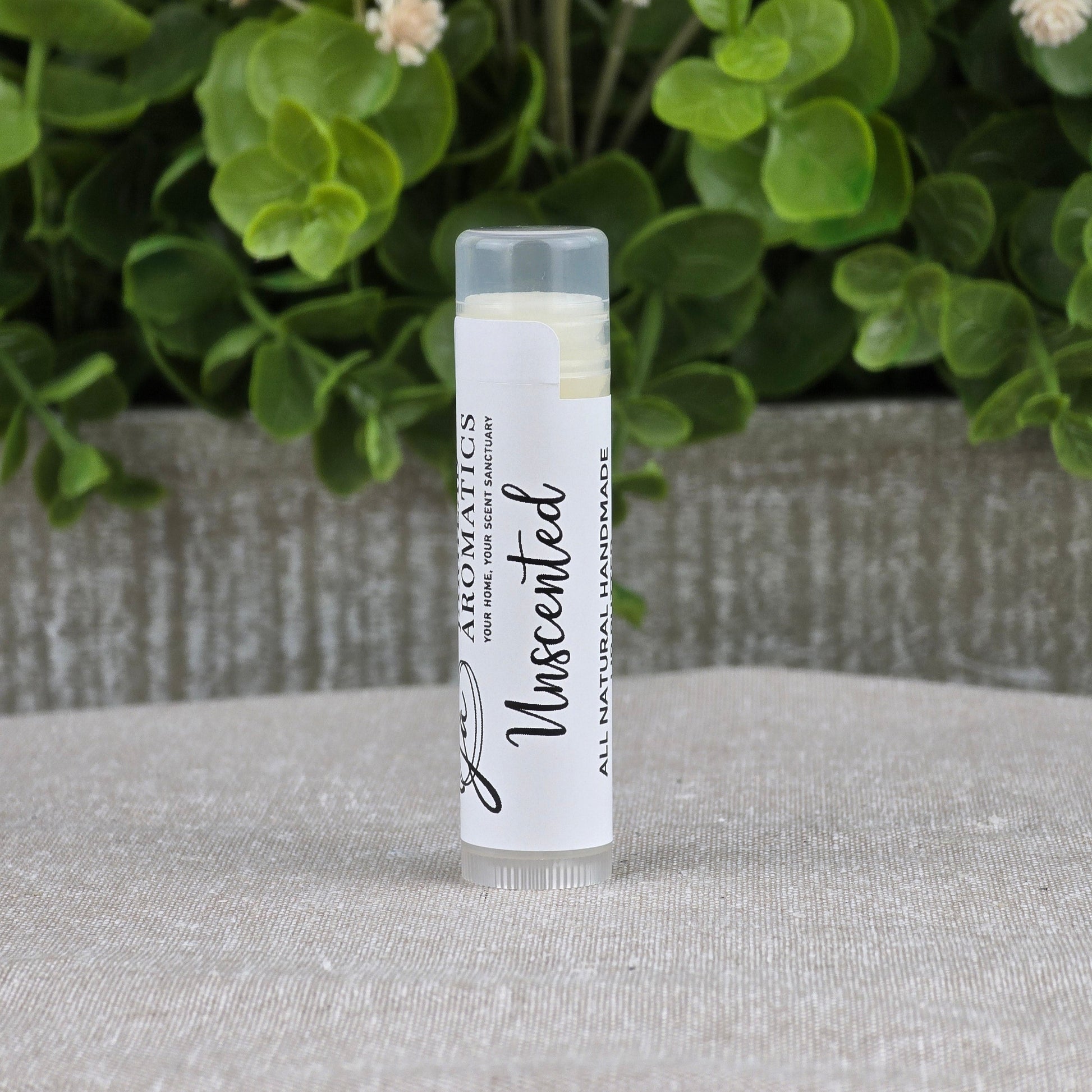 A clear tube of Jarhead Aromatics Lip Butter - All Natural stands upright on a light gray fabric, with green leafy plants and a gray planter softly blurred in the background.