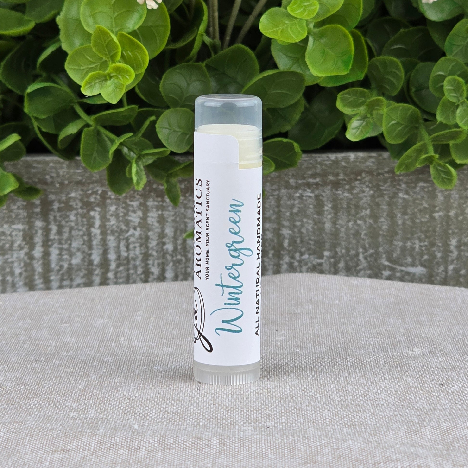 A tube of Jarhead Aromatics Lip Butter - All Natural stands upright on a textured surface, with green leafy plants and small white flowers in the background.