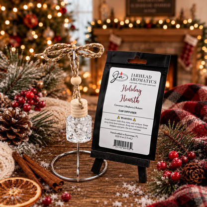 A small, clear glass Holiday Hearth Car Diffuser hanging from a metallic stand next to its packaging, set on a table decorated with aromatic cinnamon, dried citrus, and pine branches.