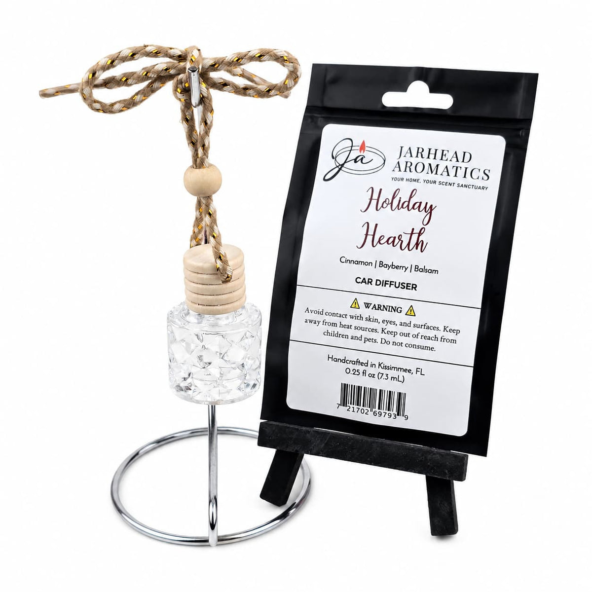 A Jarhead Aromatics Holiday Hearth Car Diffuser glass bottle hanging from a metal stand next to its black product packaging.