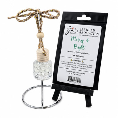 A small glass Merry & Bright Car Diffuser bottle with a wooden cap and braided hanging cord, shown next to its black retail pouch.