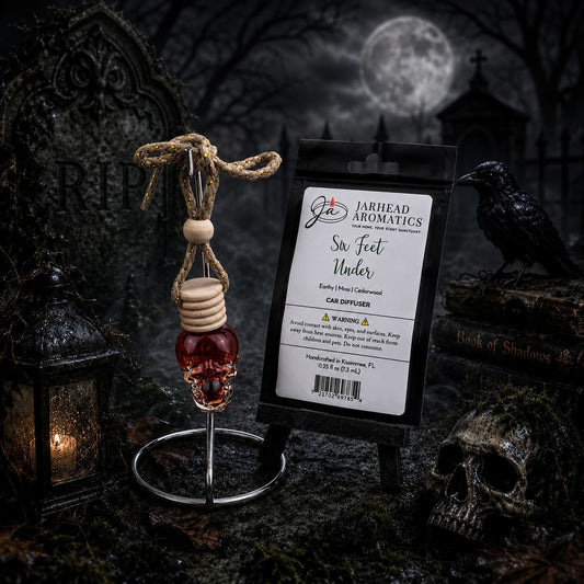 A gothic-themed display showcases the Six Feet Under Car Diffuser by Jarhead Aromatics, infused with notes of damp earth, moss, and ancient oak. The refillable air freshener is surrounded by a skull, a raven, an old lantern, and tombstones beneath a full moon, with swirling mist and eerie lighting that enhance the spooky atmosphere.