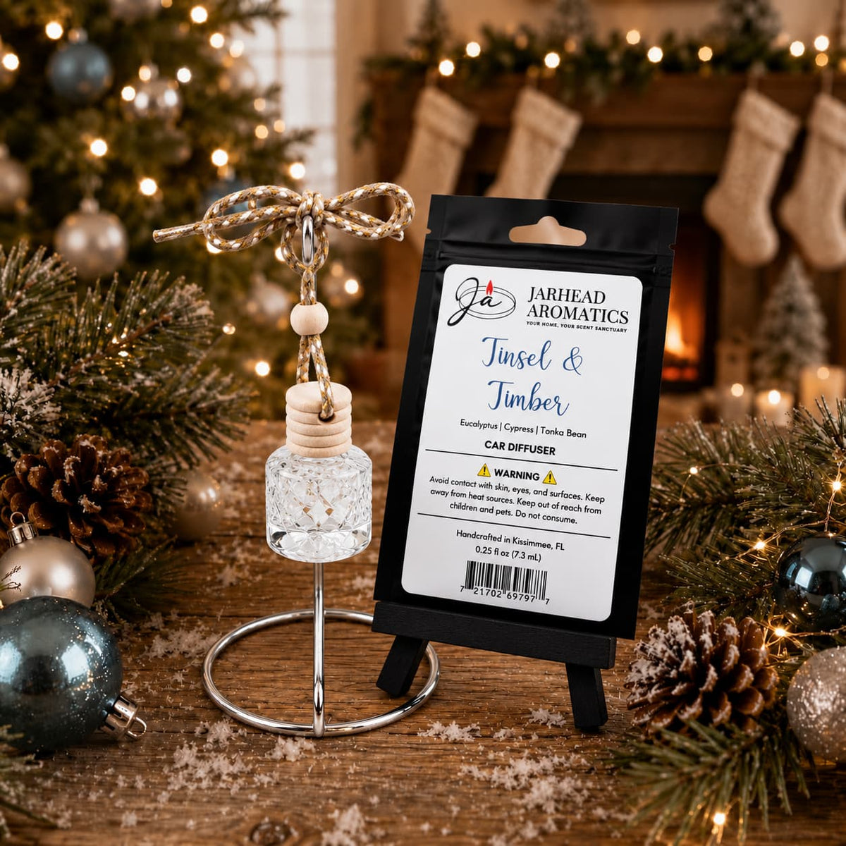 A Jarhead Aromatics Tinsel & Timber Car Diffuser with a wooden cap and hanging cord stands next to its packaging on a wooden table decorated with pine branches and ornaments, with a fireplace in the background.
