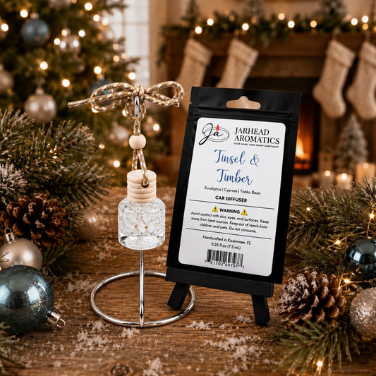 A Jarhead Aromatics Tinsel & Timber Car Diffuser with a wooden cap and hanging cord stands next to its packaging on a wooden table decorated with pine branches and ornaments, with a fireplace in the background.